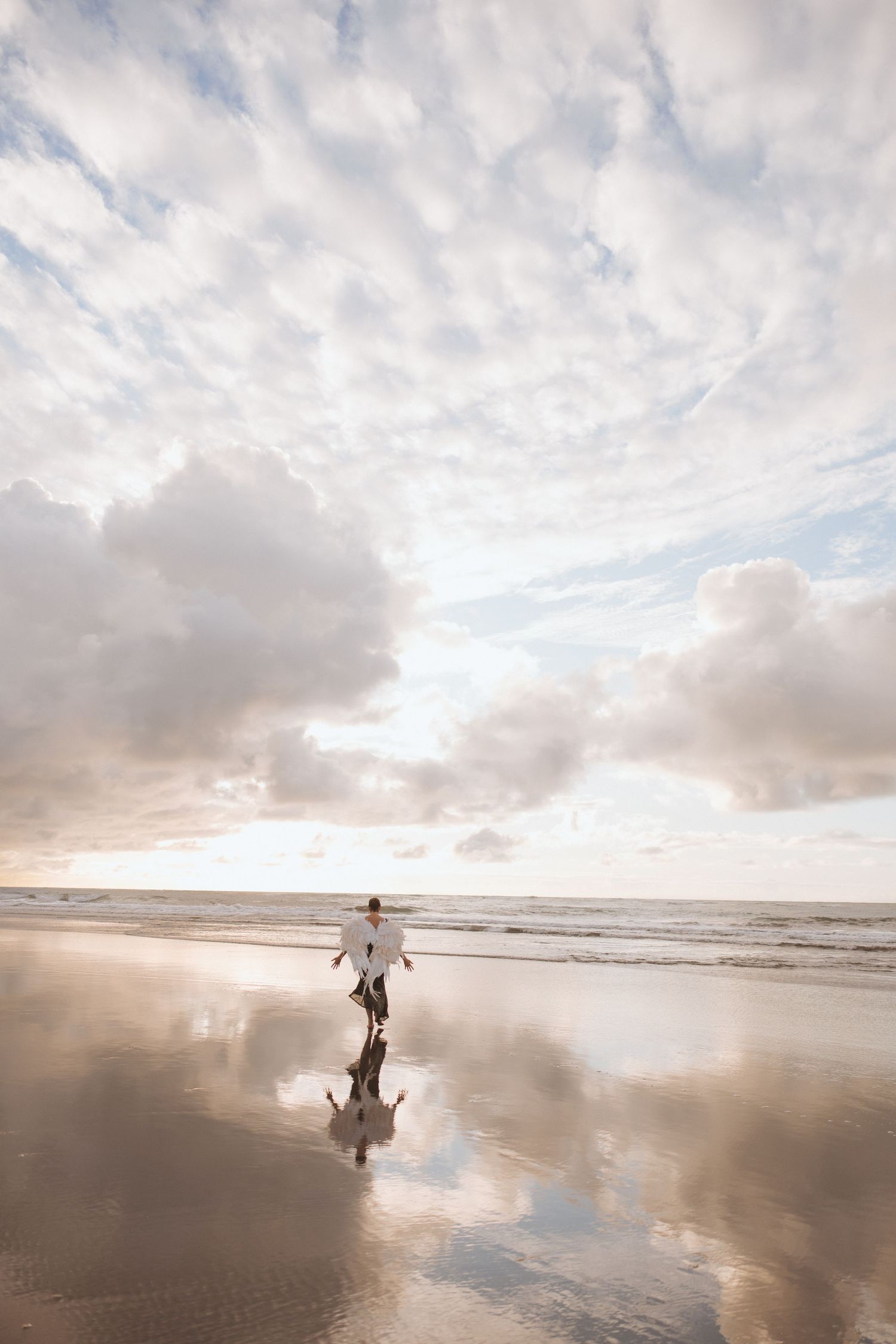 Person in white dress walking on reflective wet beach with dramatic cloud-filled sky at sunset in New Zealand.