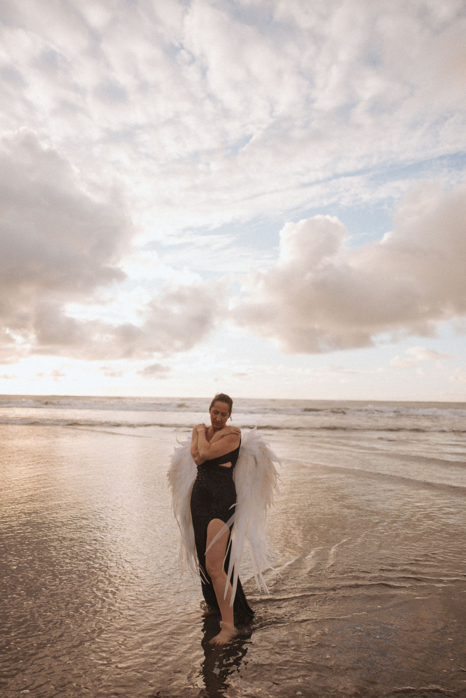 Woman in black dress with white angel wings standing in ocean surf at golden sunset in New Zealand.