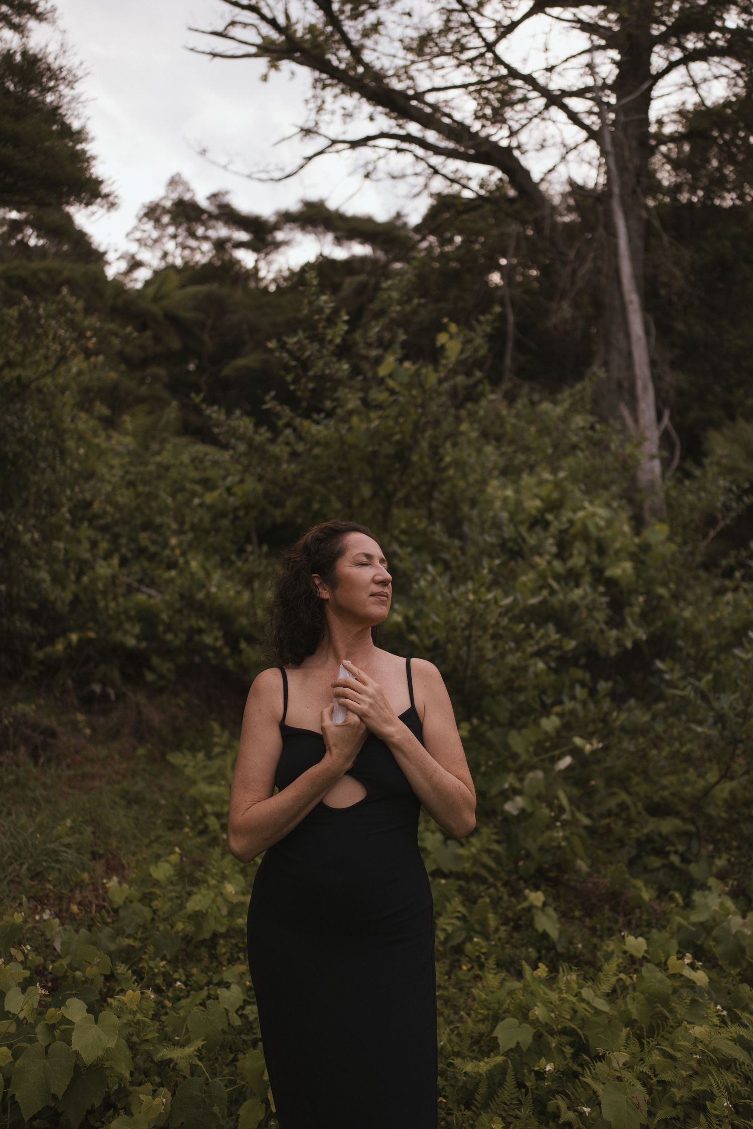Woman in black slip dress standing peacefully in lush green forest with eyes closed in New Zealand.