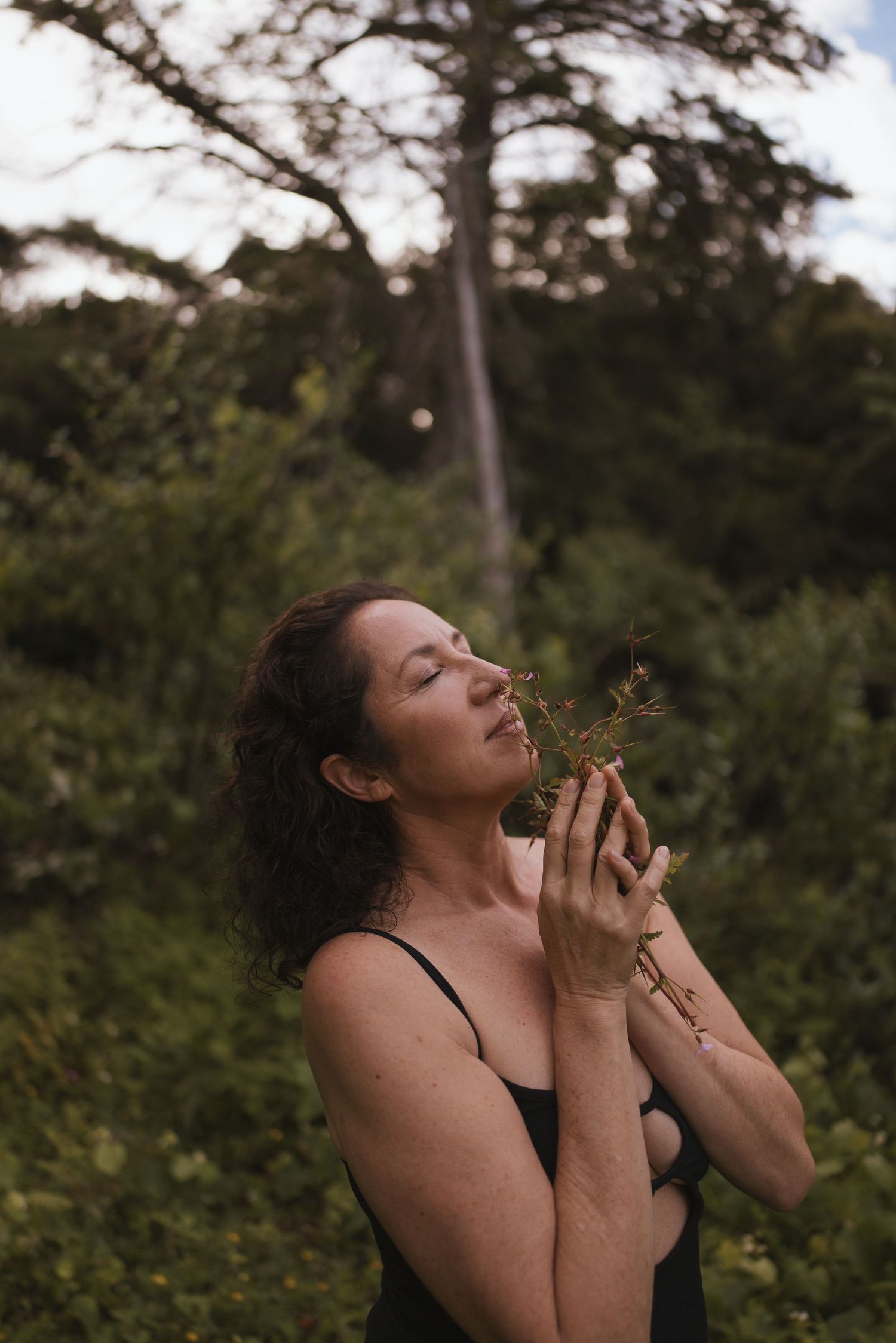 A woman with closed eyes holds a small plant to her chest in a lush green forest setting in New Zealand.