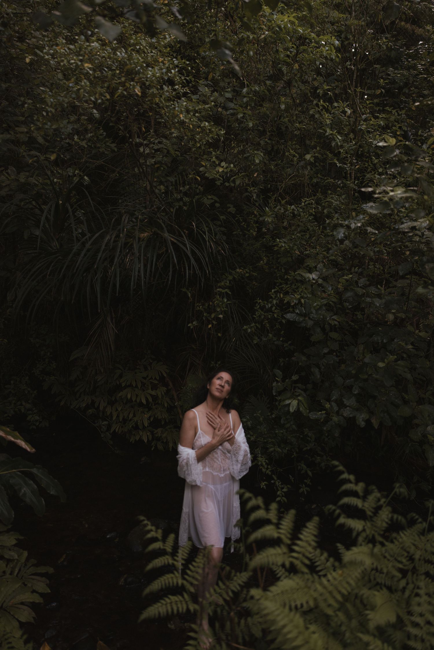A woman in a white sheer dress stands peacefully among dense green ferns and tropical foliage in New Zealand.