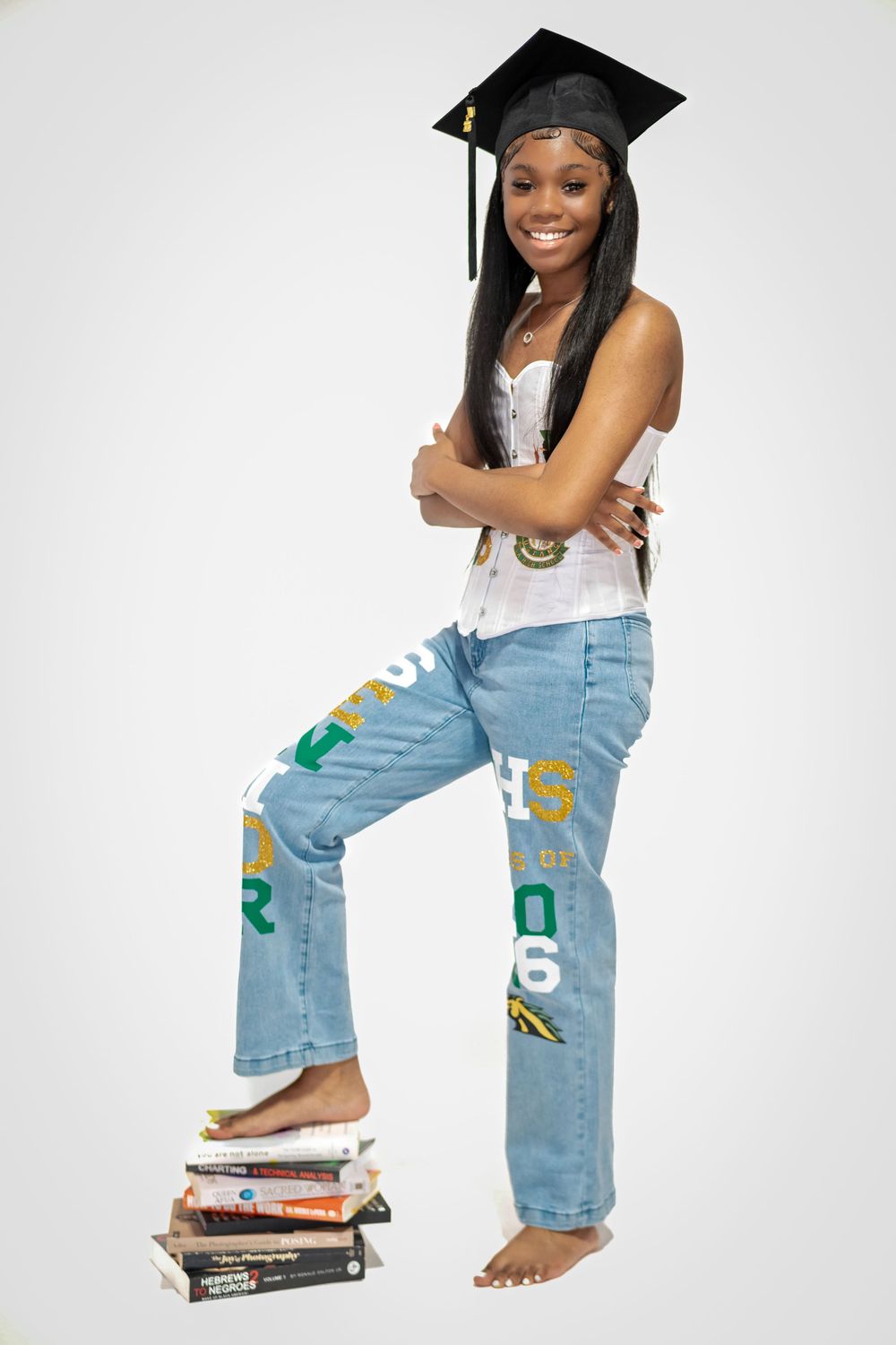 Graduate in decorated jeans and cap sitting on stack of books.