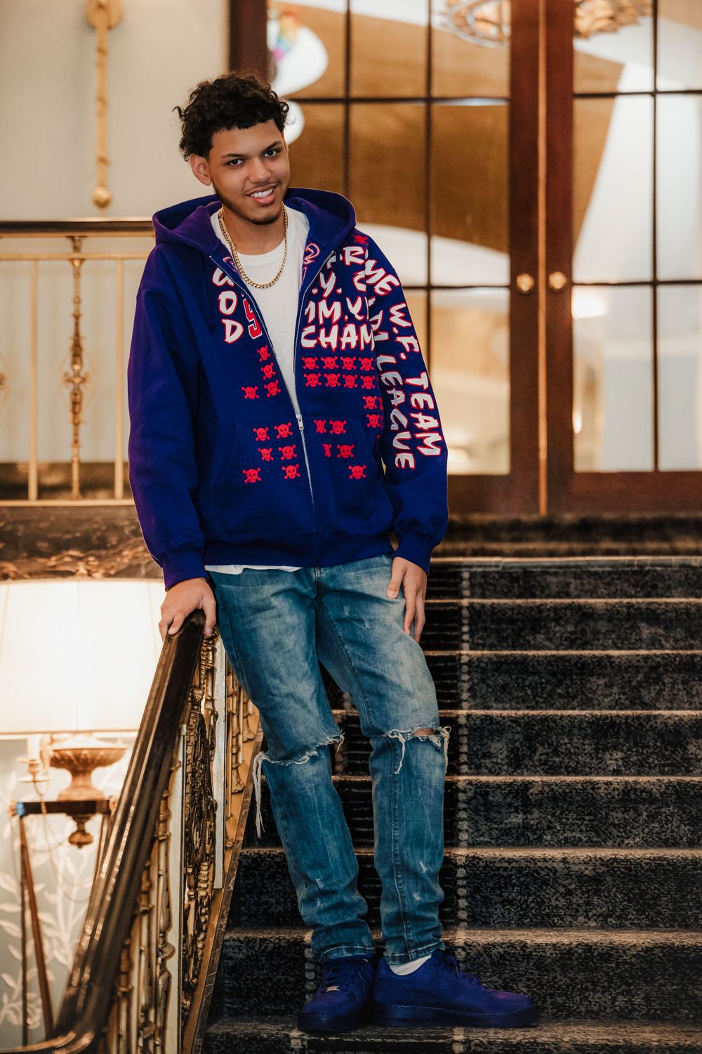 Person poses on stone steps wearing blue varsity jacket and ripped jeans.