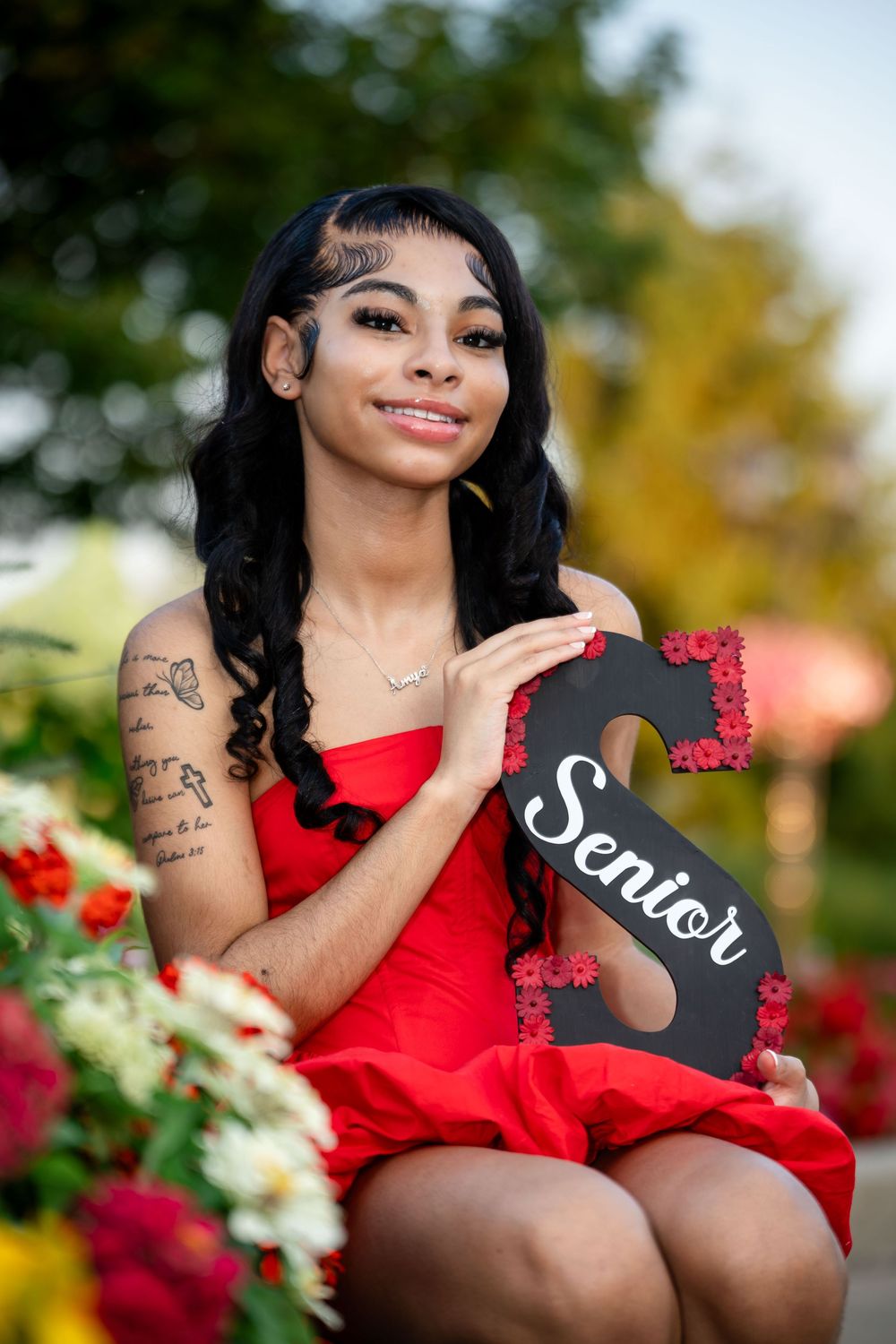 Graduate in red dress posing with senior sign among flowers.