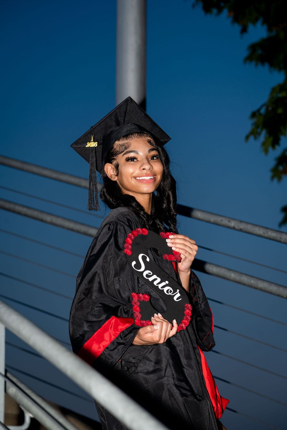 Graduate in black cap and gown with red accents against blue sky background.