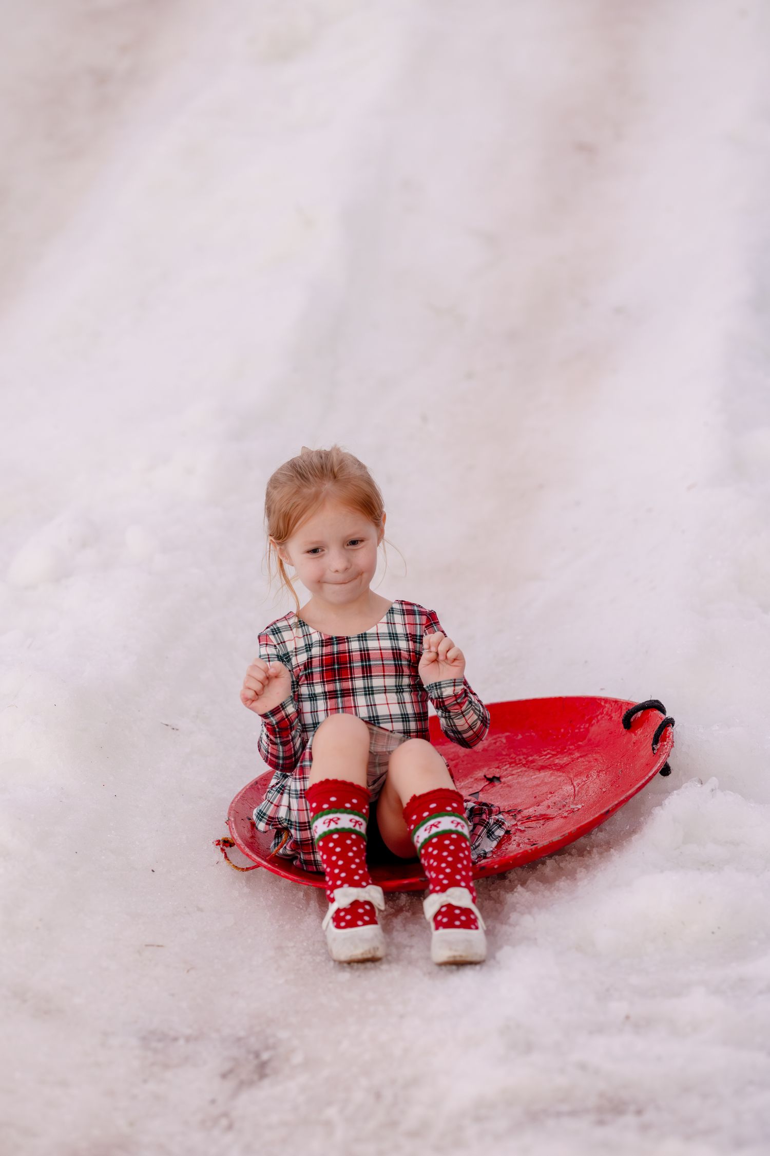A child in a plaid shirt sits in fresh snow with a bright red sled, creating a festive winter scene.