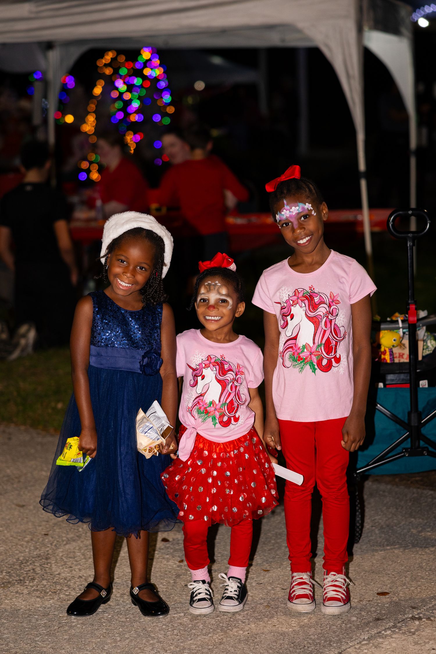 Three children in festive outfits sitting together at a nighttime outdoor event with colorful lights.