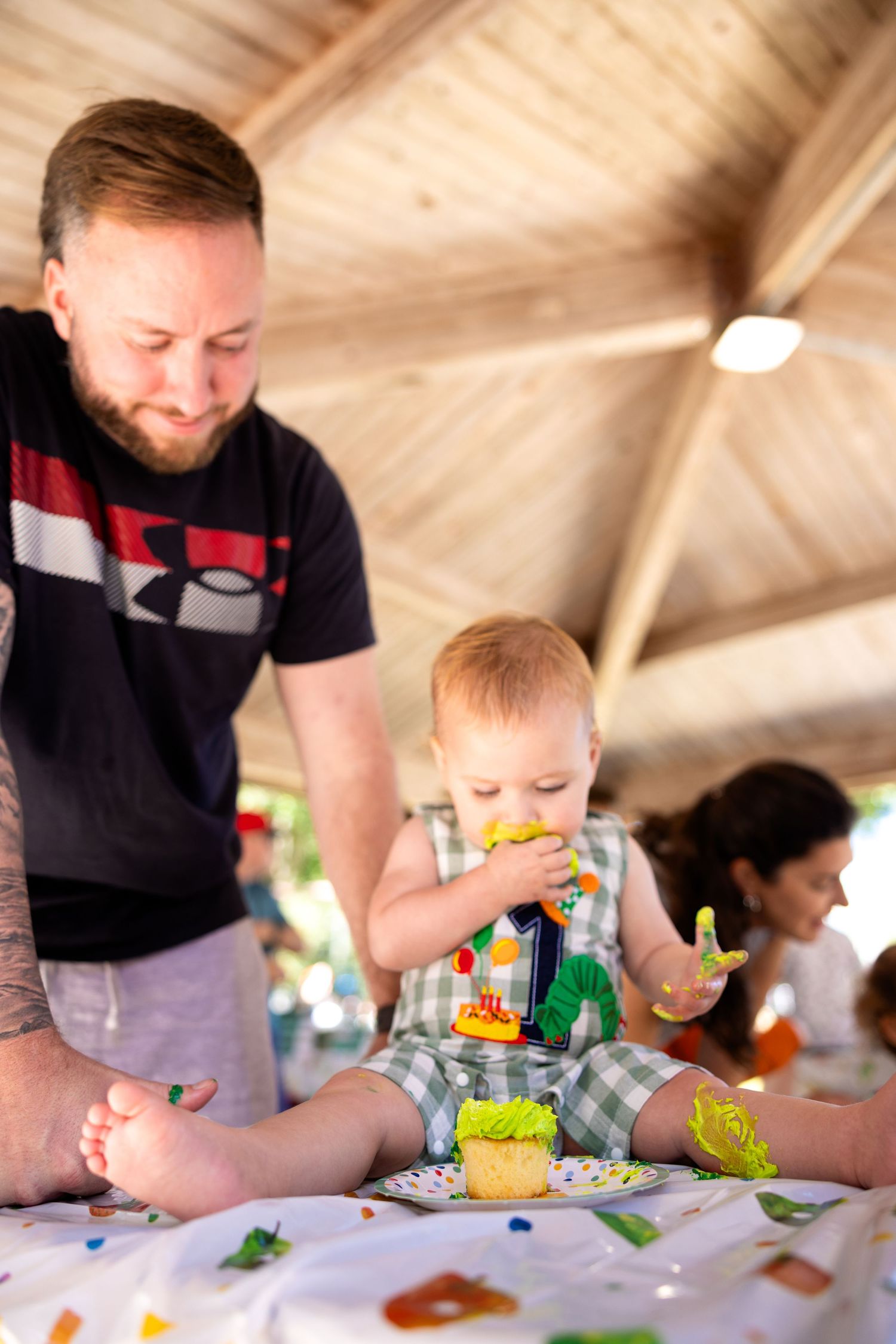 A toddler in plaid shorts plays with toys at a table while an adult watches during what appears to be a party or gathering.