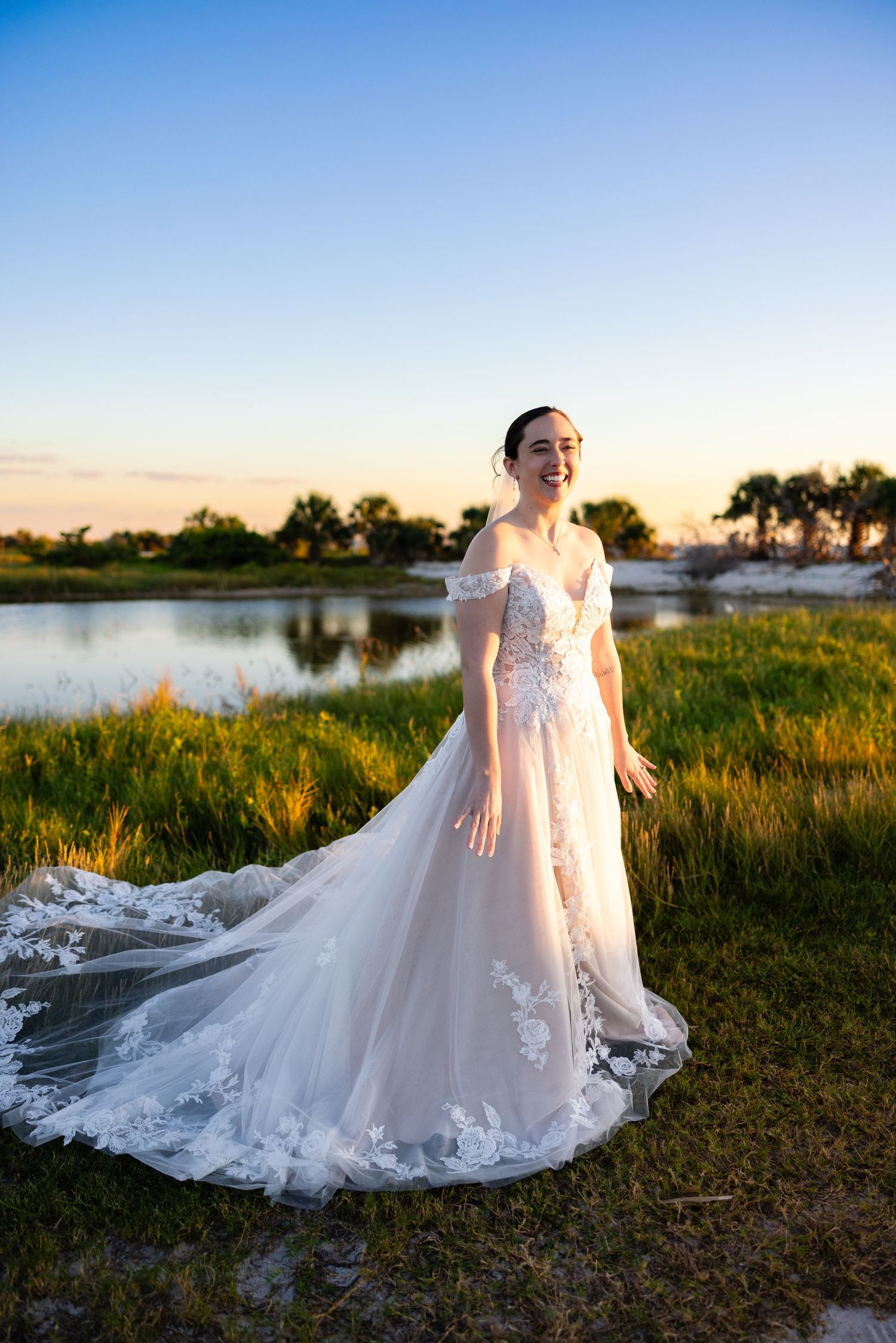 A stunning bridal portrait session at sunset showcases an elegant lace wedding gown with flowing train by a lake.