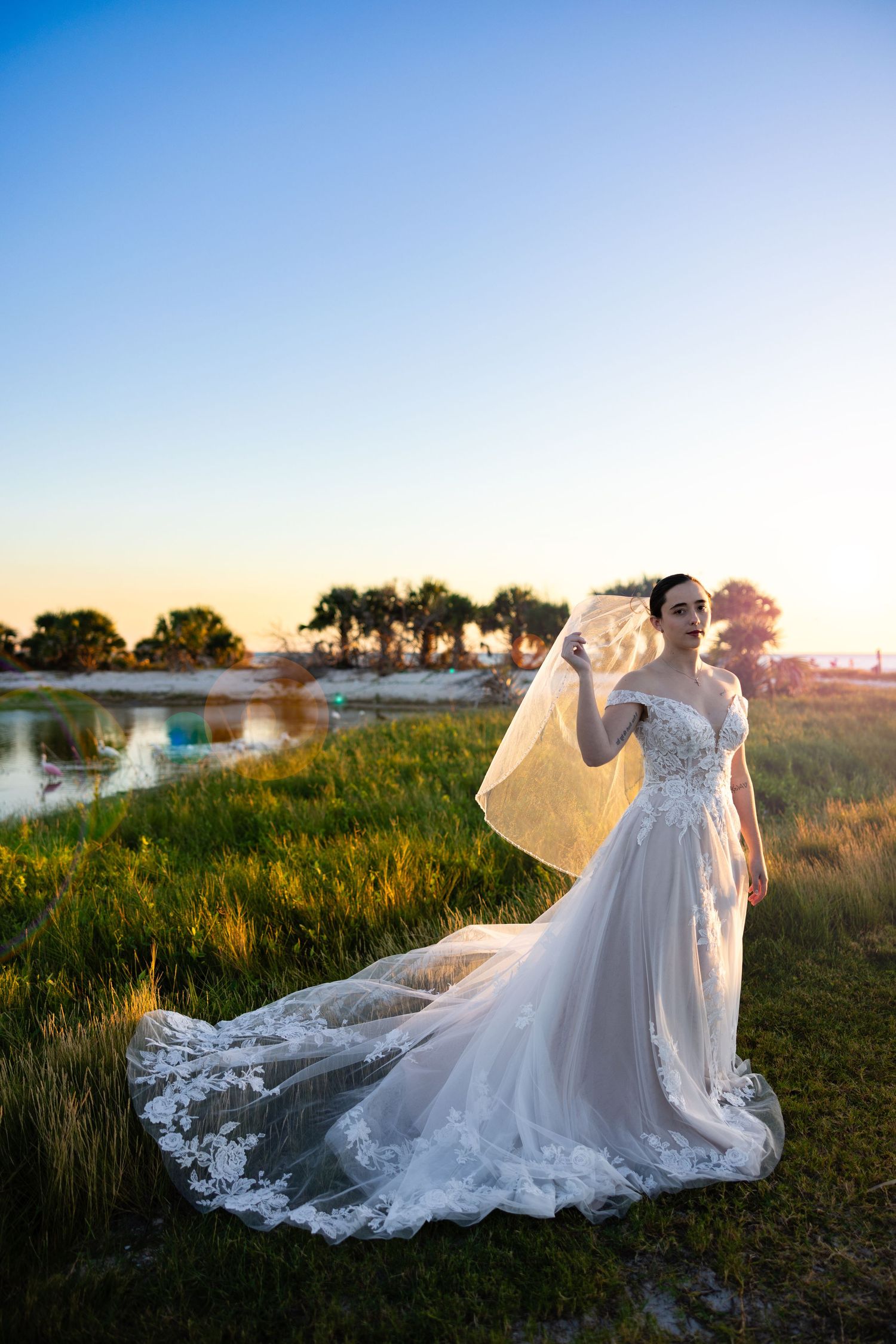 A bride in a flowing lace wedding gown with long train poses at sunset near a waterfront with palm trees in the background.
