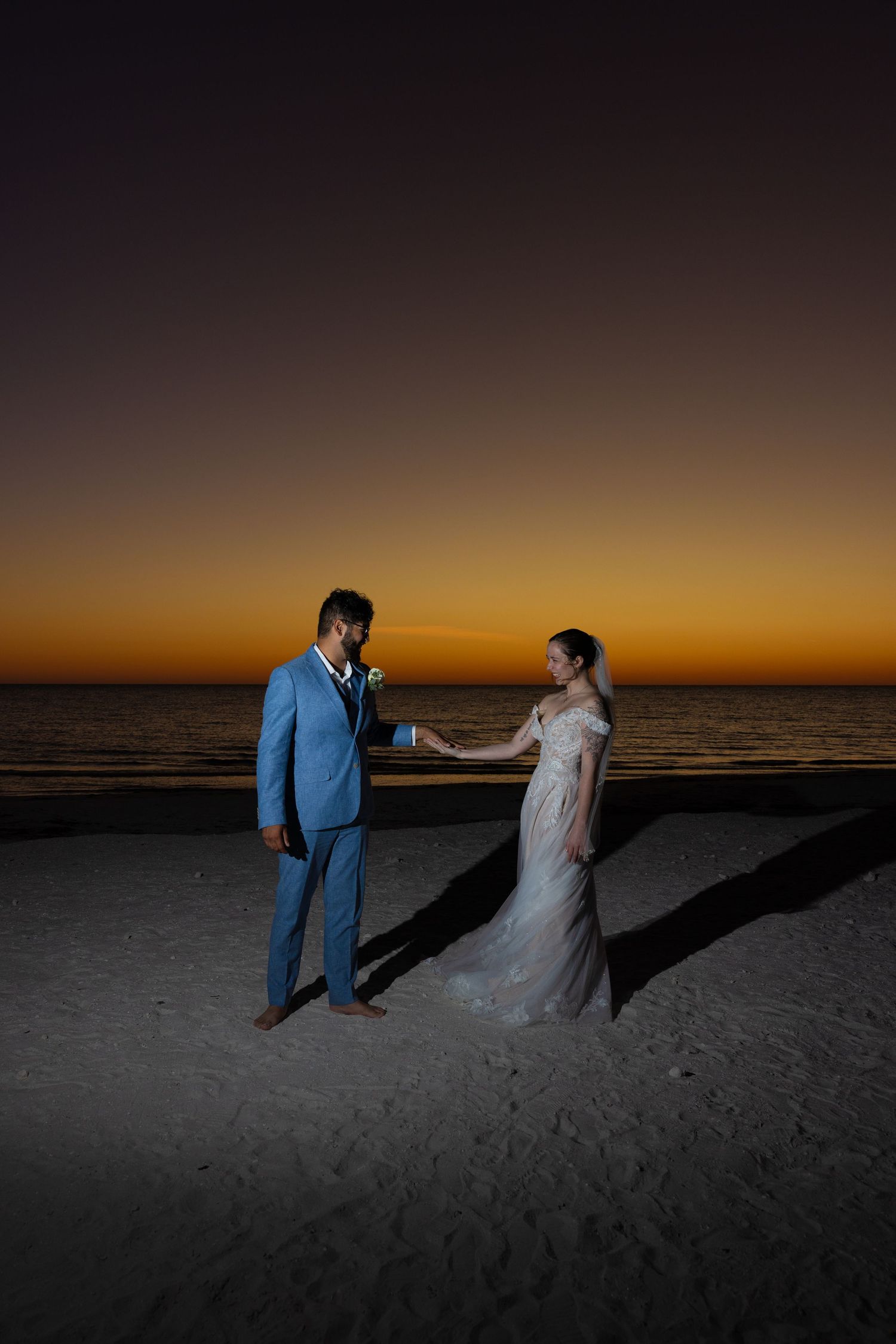 A couple in wedding attire holds hands on a sandy beach at sunset with a dramatic orange sky in the background.