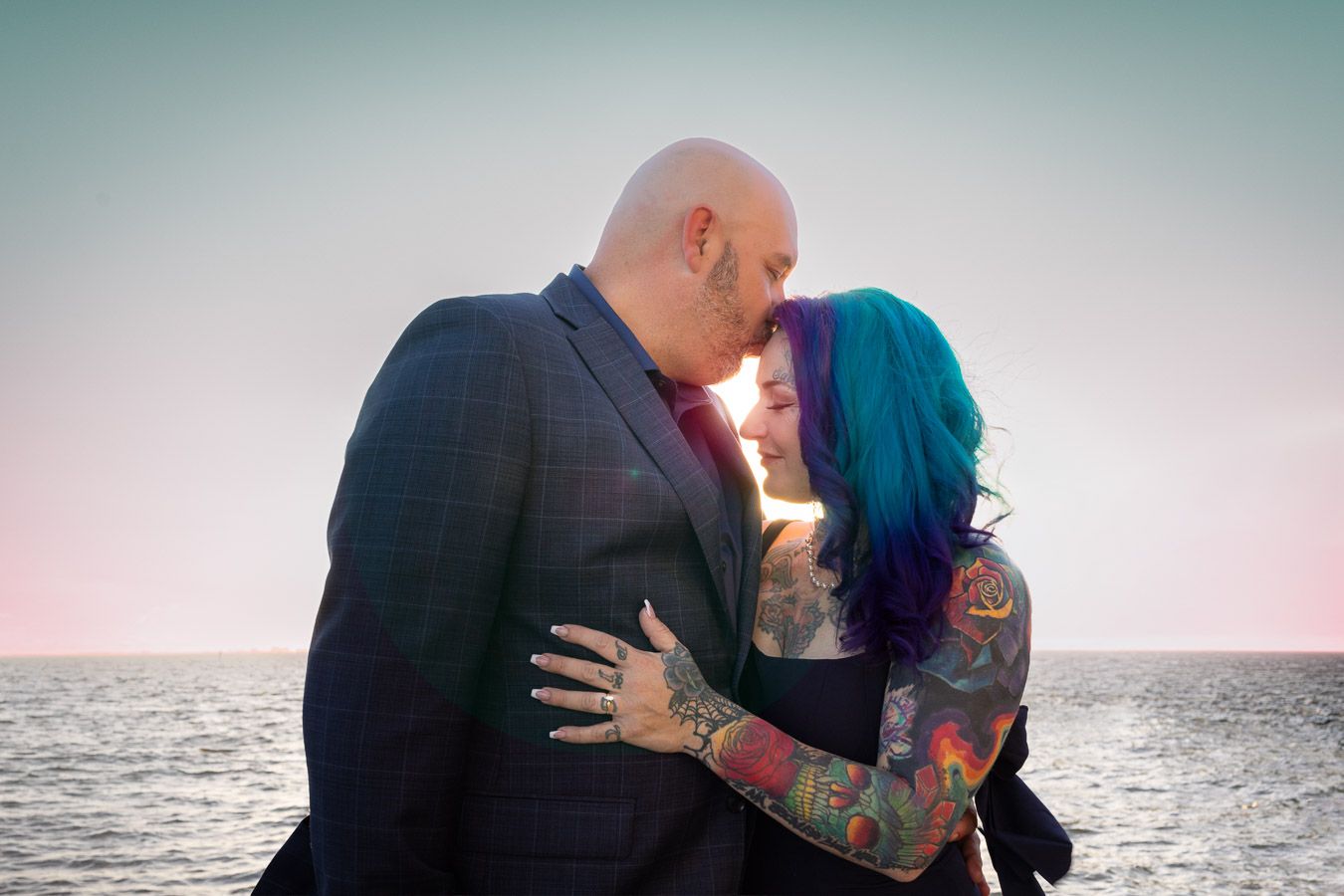 A couple embraces in a romantic kiss at sunset by the ocean, with one person sporting vibrant blue hair and tattoos.