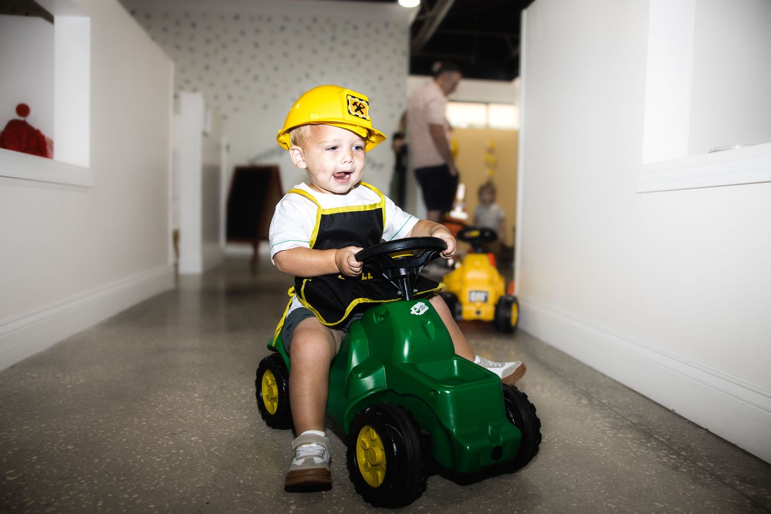 A child wearing a yellow hat rides a green John Deere toy tractor through a bright white hallway.