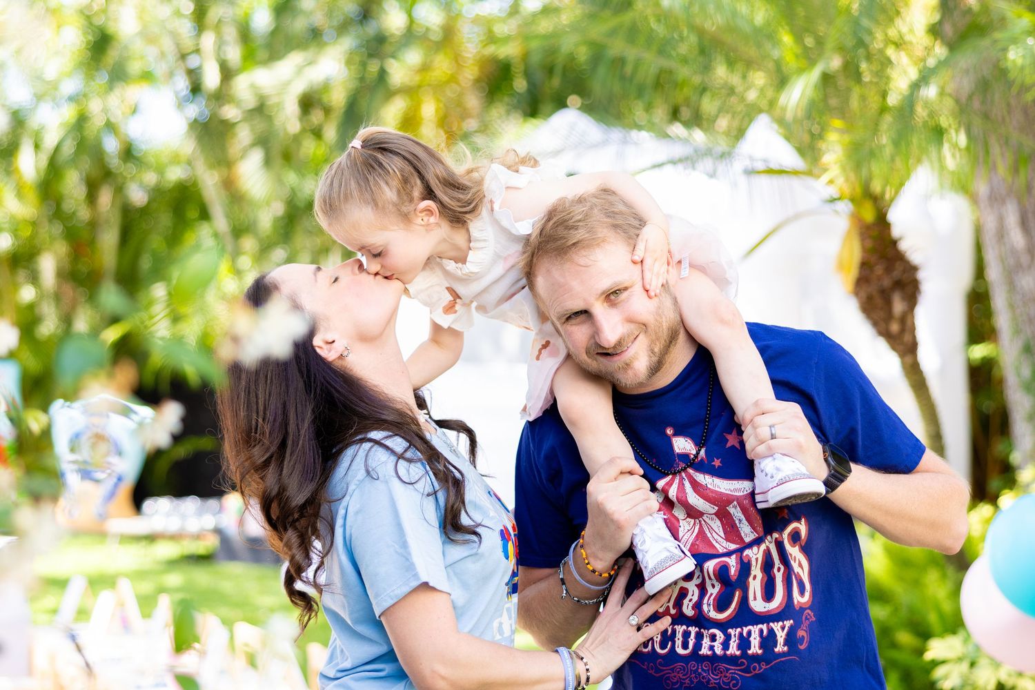 Family of four enjoying a playful moment together outdoors in a sunny garden setting with trees in the background.