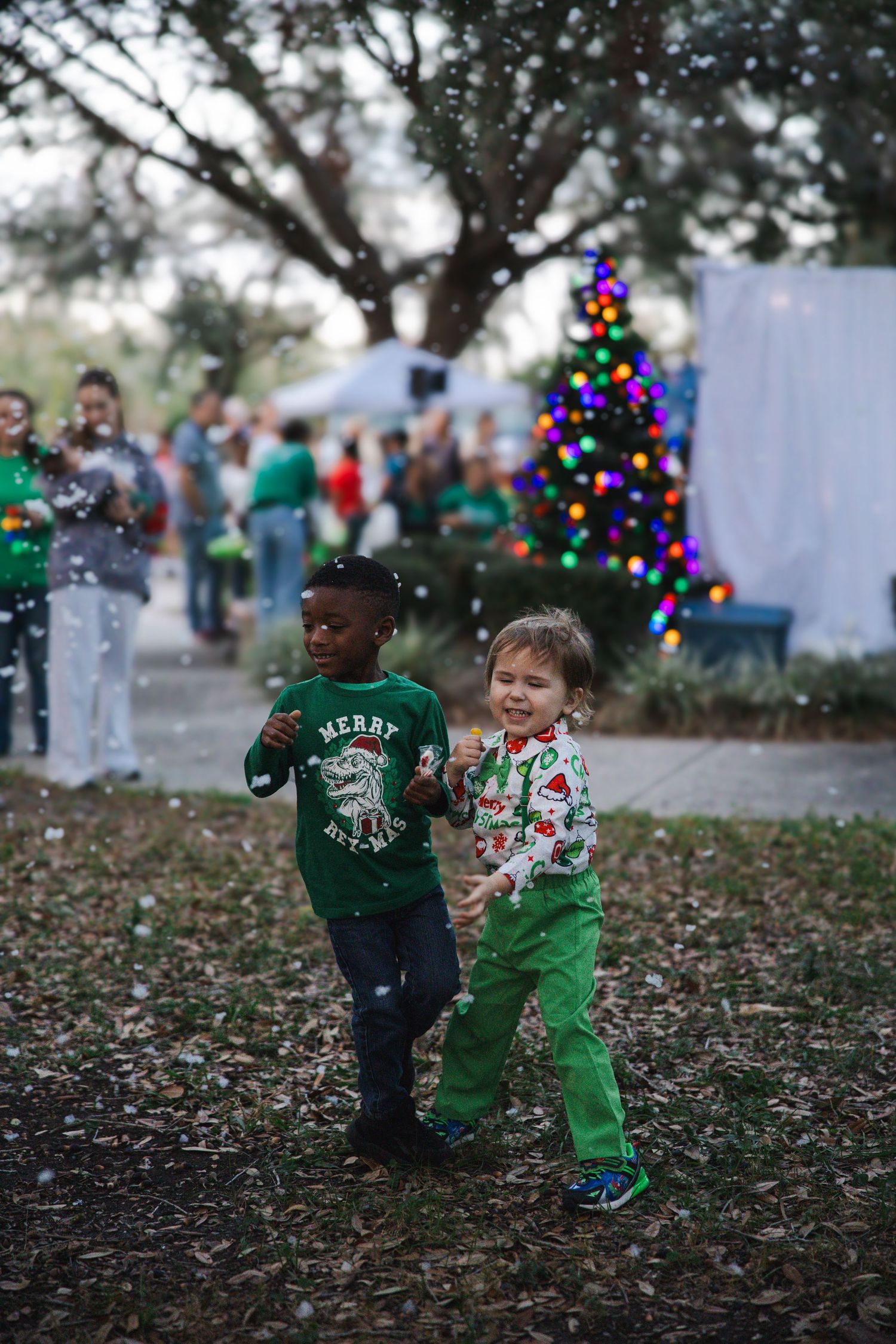 Two children in festive sweaters play near a lit Christmas tree outdoors.
