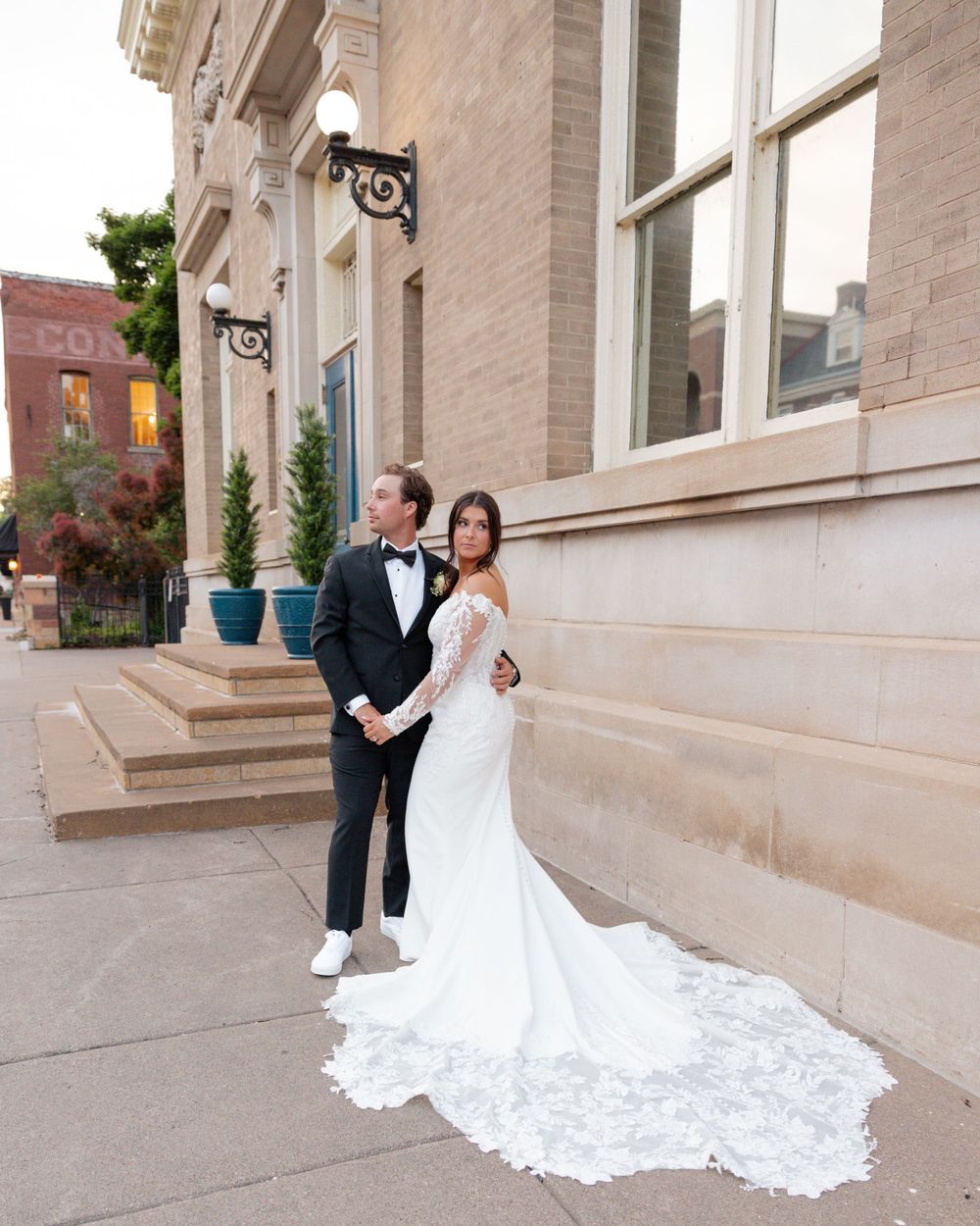 A formal couple poses outside a classic stone building, showcasing a flowing white wedding dress train in Stillwater, MN.