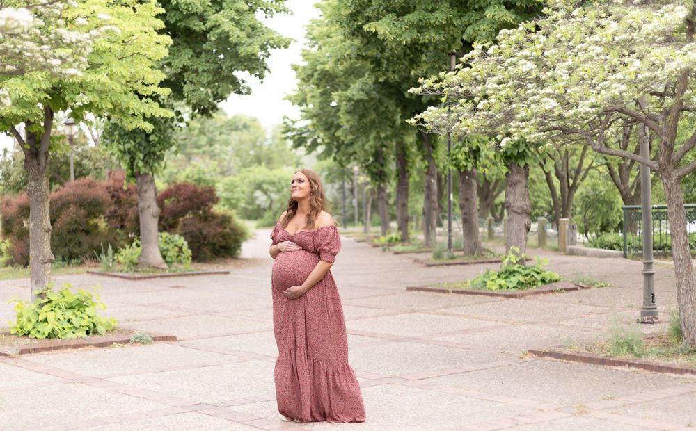 A maternity portrait in a pink dress surrounded by flowering trees in a park setting.