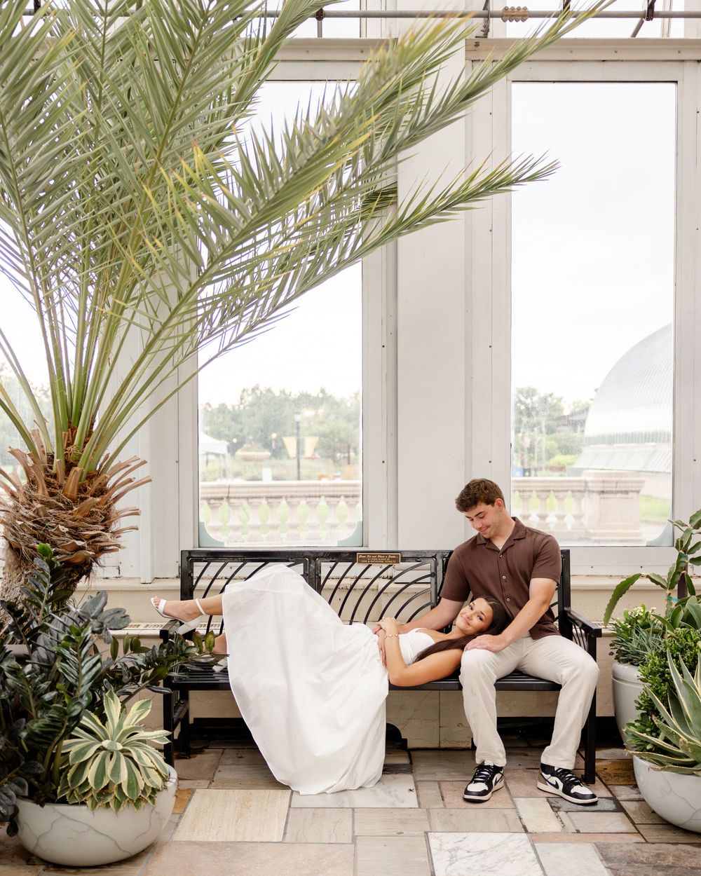 A romantic couple in wedding attire sits together in a modern greenhouse setting with palm plants and white planters.