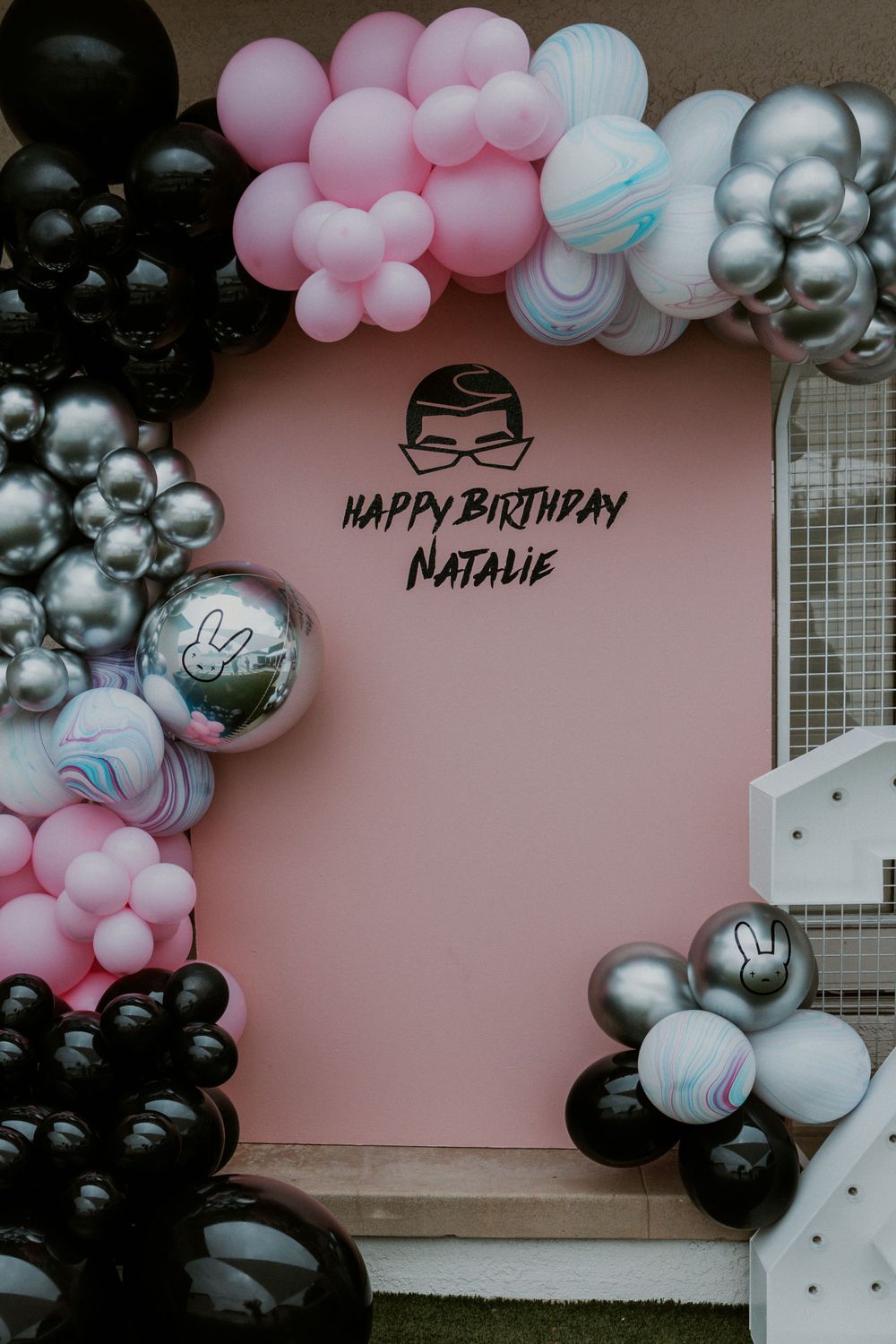 Birthday celebration backdrop with black, pink and silver balloon arch against a pink wall.
