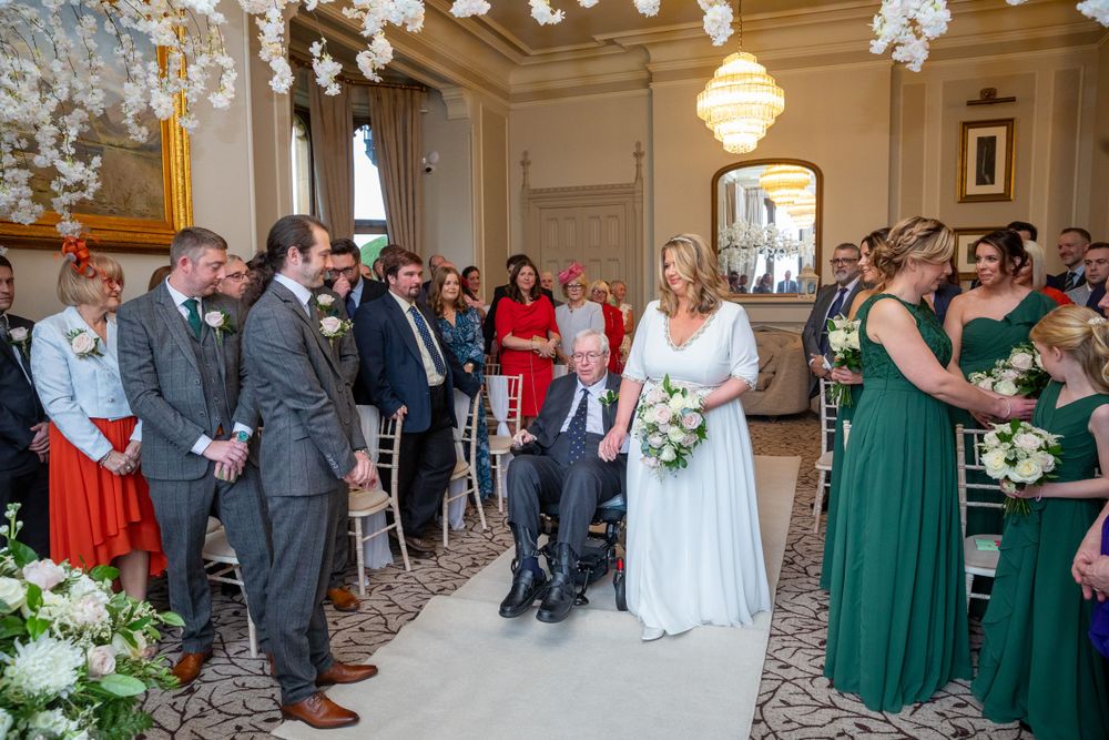 Bride walks down aisle with father in wheelchair at elegant wedding ceremony.