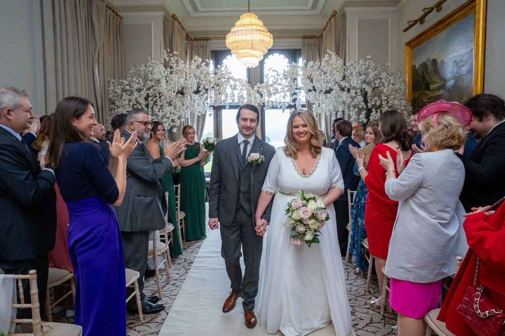 Newlyweds walk hand in hand down aisle past cheering guests in ornate wedding venue.