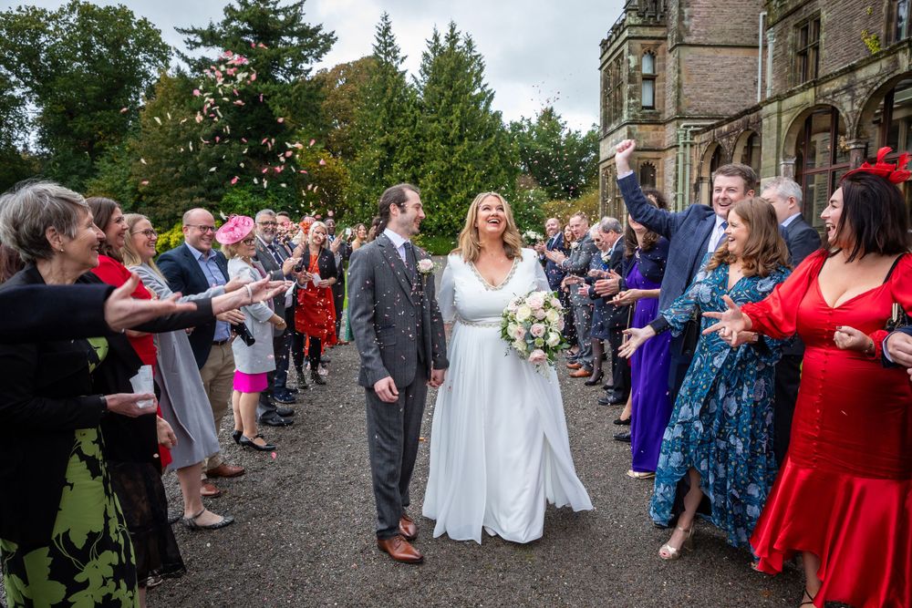 Smiling newlyweds walk through confetti shower surrounded by celebrating wedding guests.