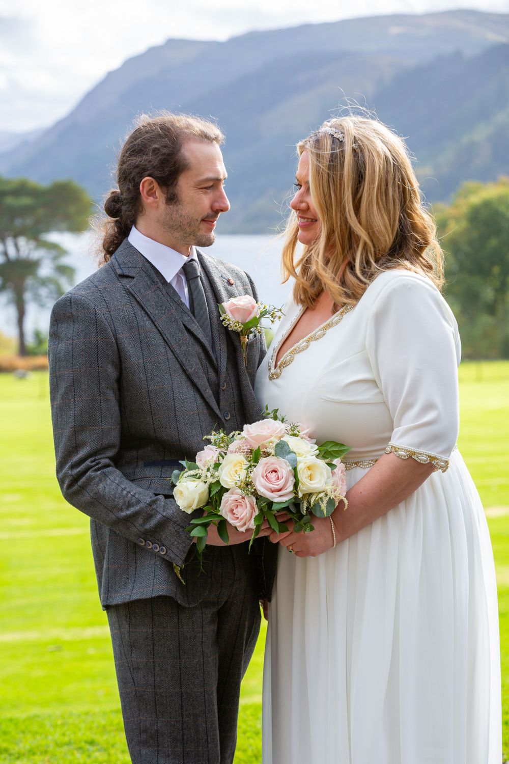 Bride and groom share a tender moment outdoors with mountain lake backdrop, bride holding pink rose bouquet.
