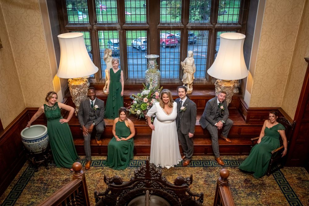 Wedding party poses on grand staircase, bridesmaids in emerald green dresses flanking bride and groom.