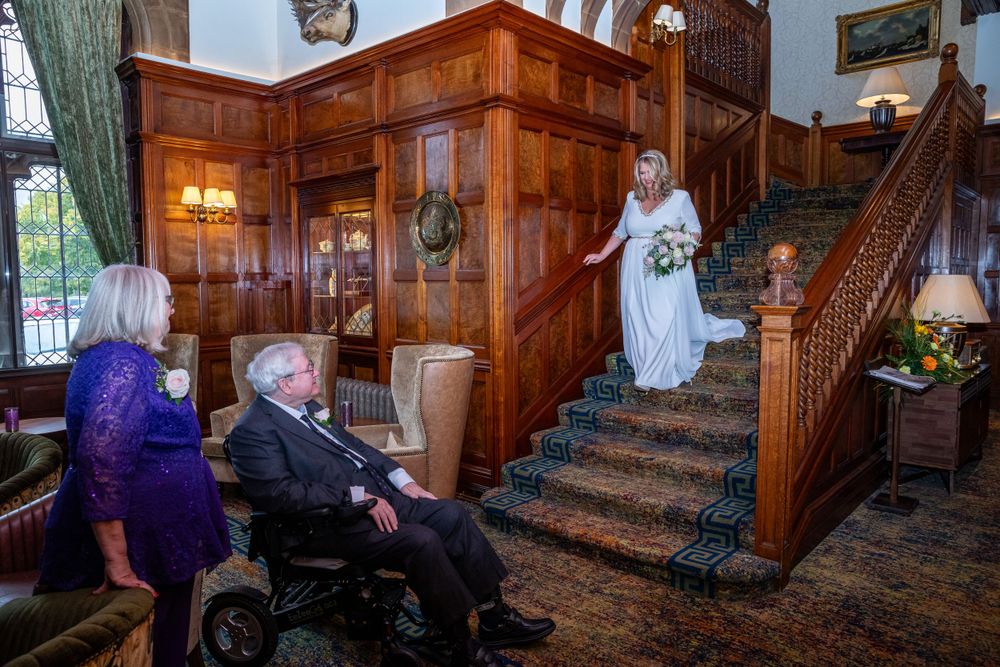 Bride descends grand wooden staircase in elegant manor house as family watches.