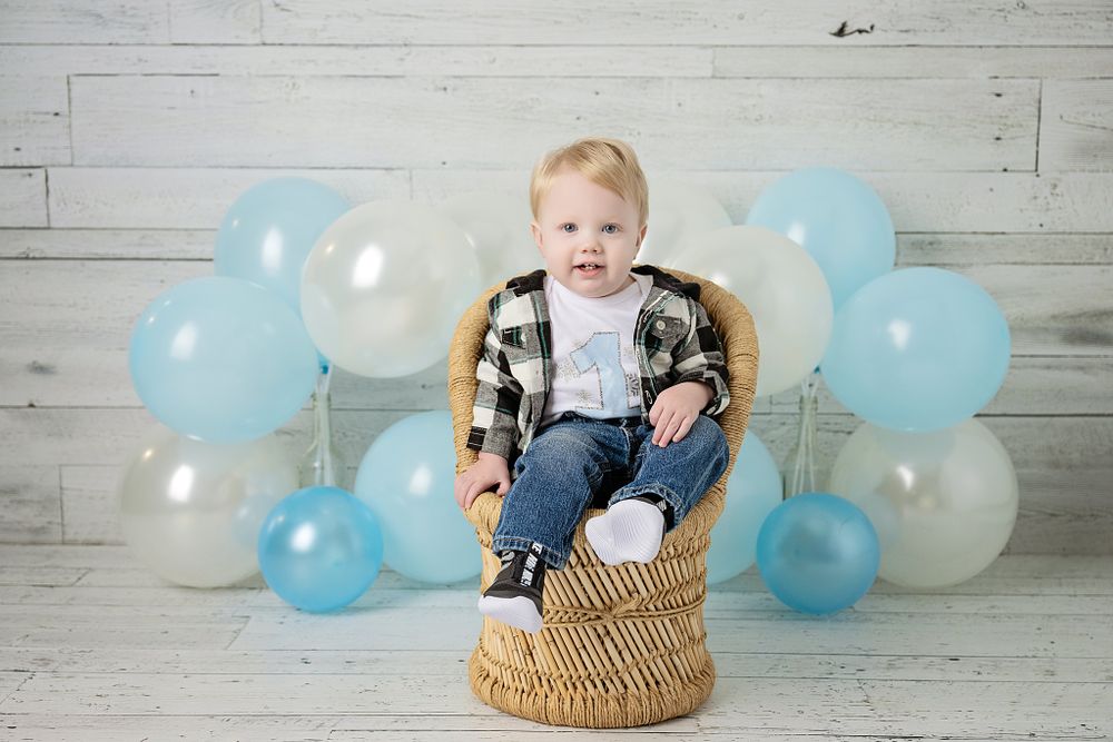 First birthday photo with blue balloons, rattan chair and ONE letters against white wood wall.