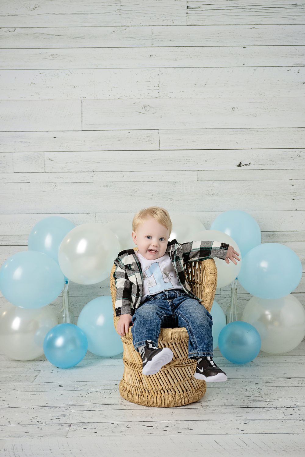 First birthday photo with plaid shirt, rattan chair, and blue balloon backdrop.