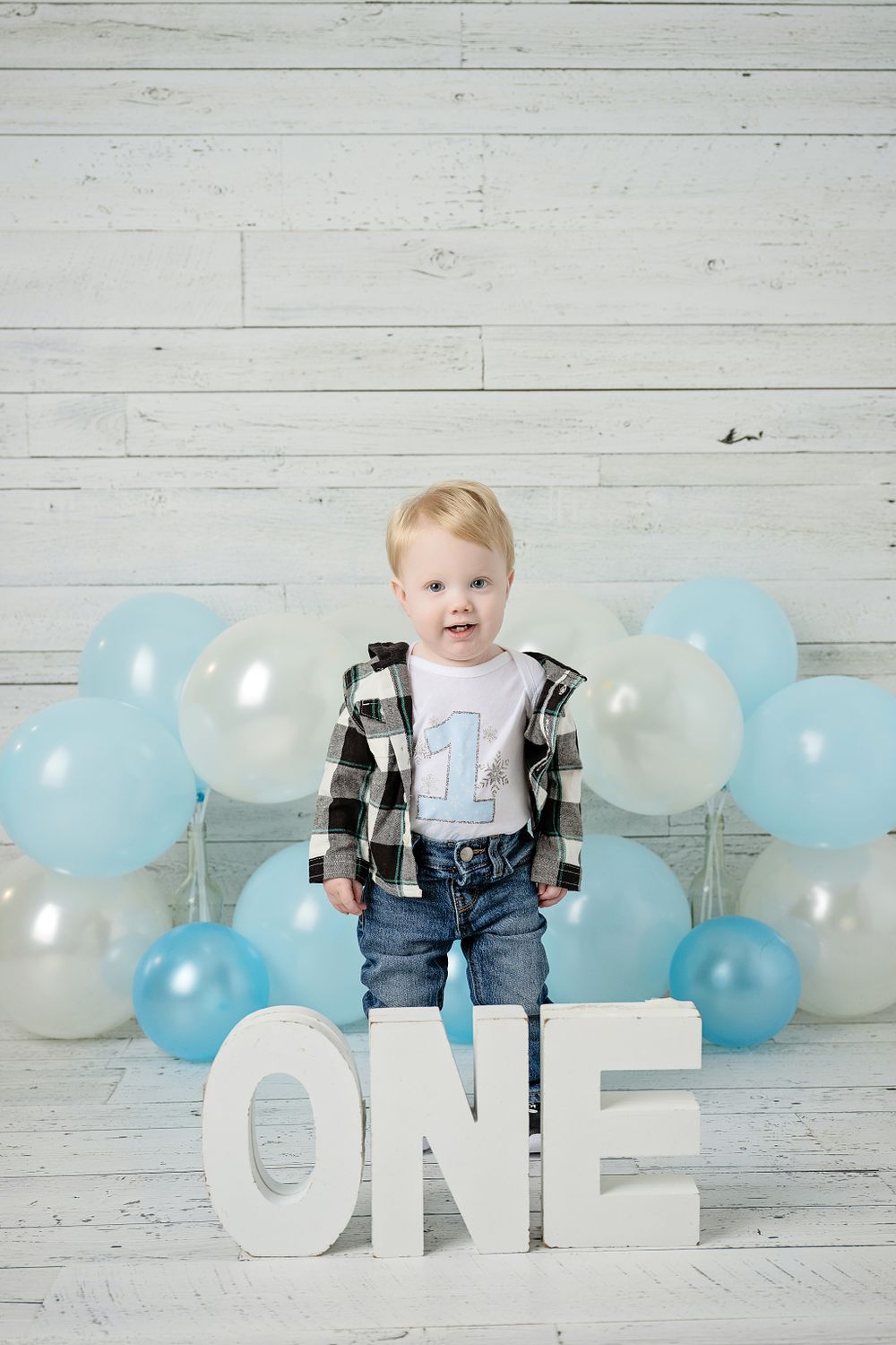 First birthday scene with white ONE letters, blue balloons, and white wood background.