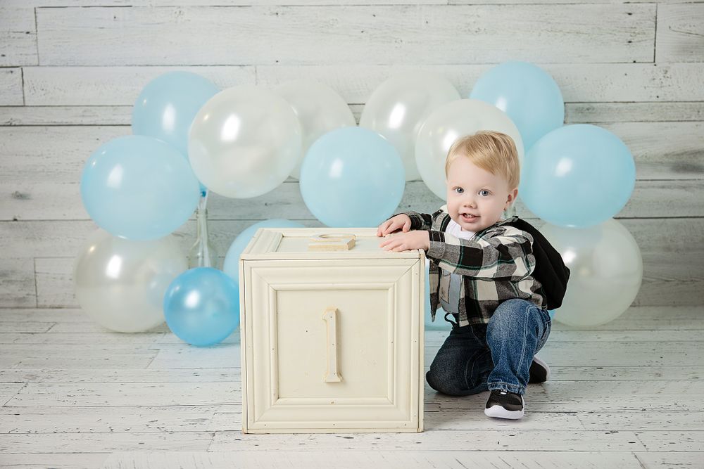 First birthday photo featuring wooden block, plaid shirt, and blue balloon decorations.