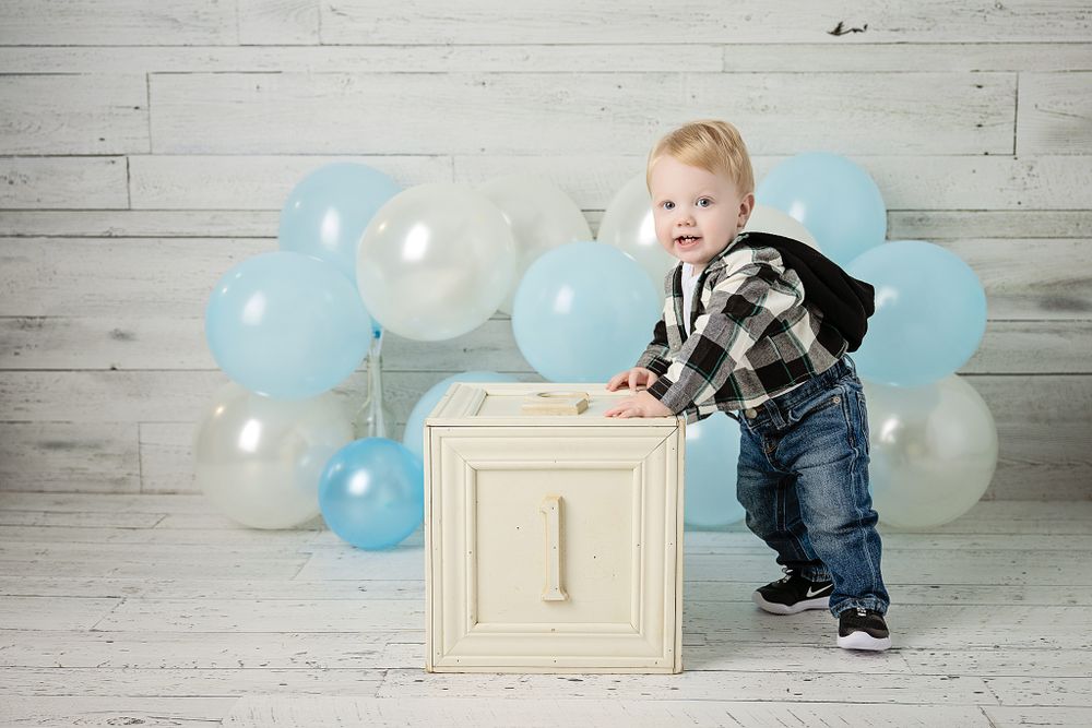 First birthday photo setup with white wooden block, plaid shirt, and blue and white balloon backdrop.