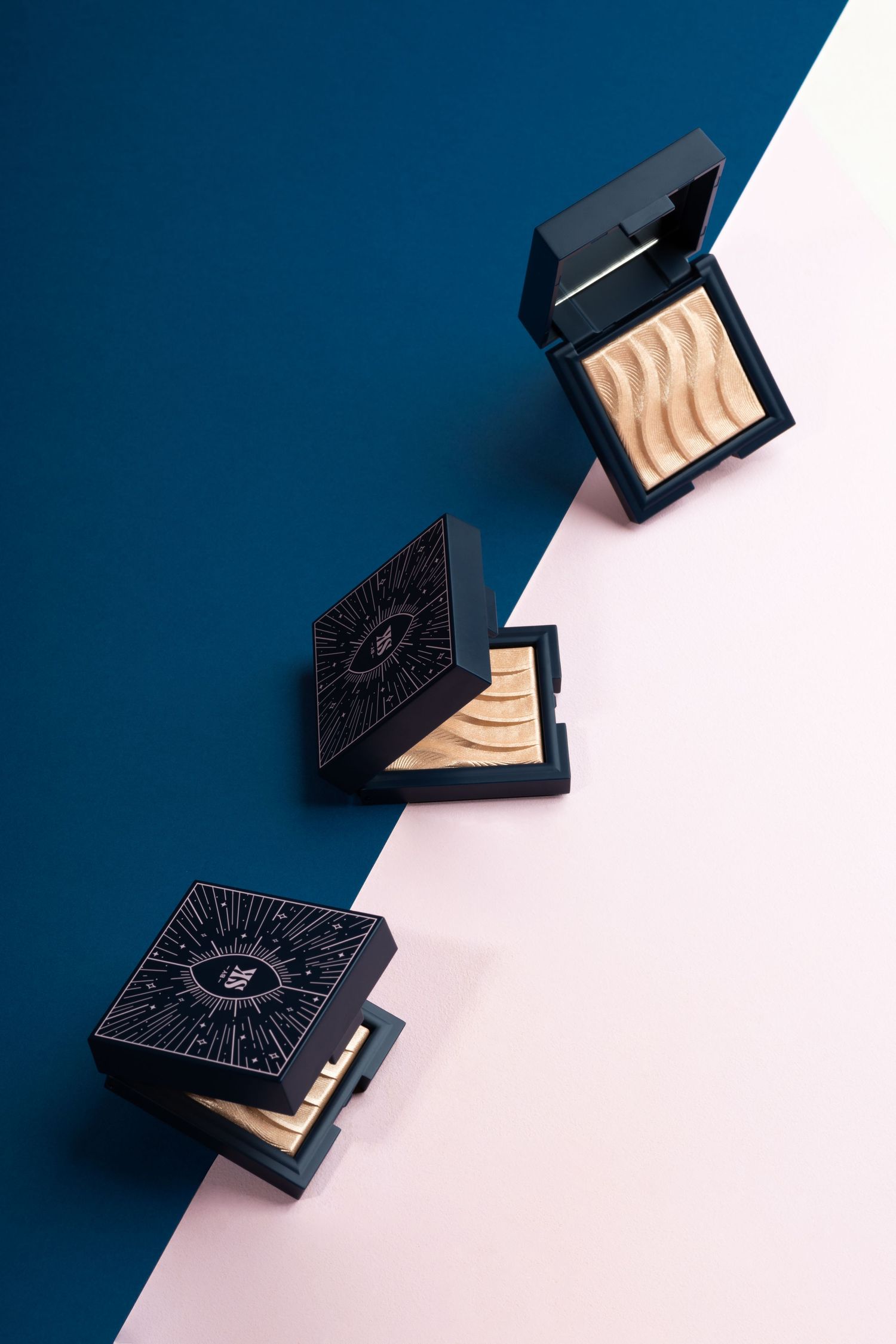 Three black makeup compacts with geometric designs displaying highlighter powders on navy blue and baby pink split background.