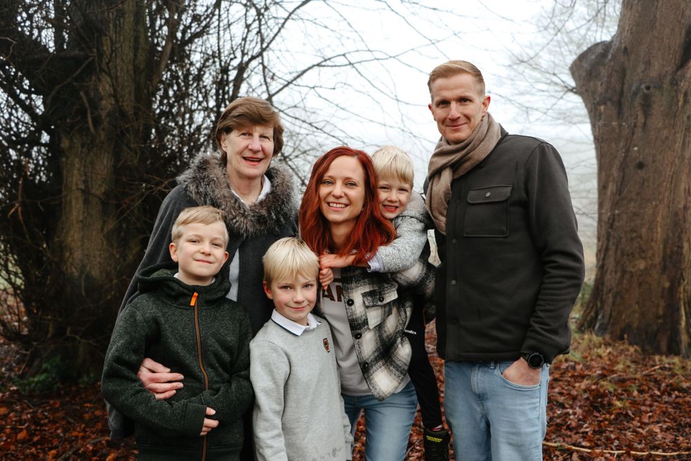 A group poses together outdoors on fallen autumn leaves between tall trees in a forest setting.
