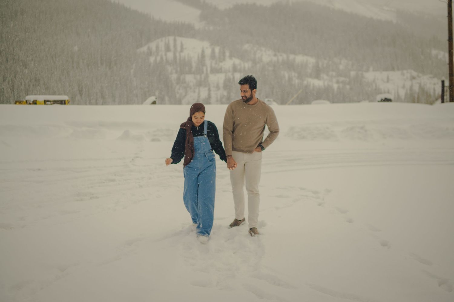 A couple walking together in a snowy winter landscape with mountains in the background.
