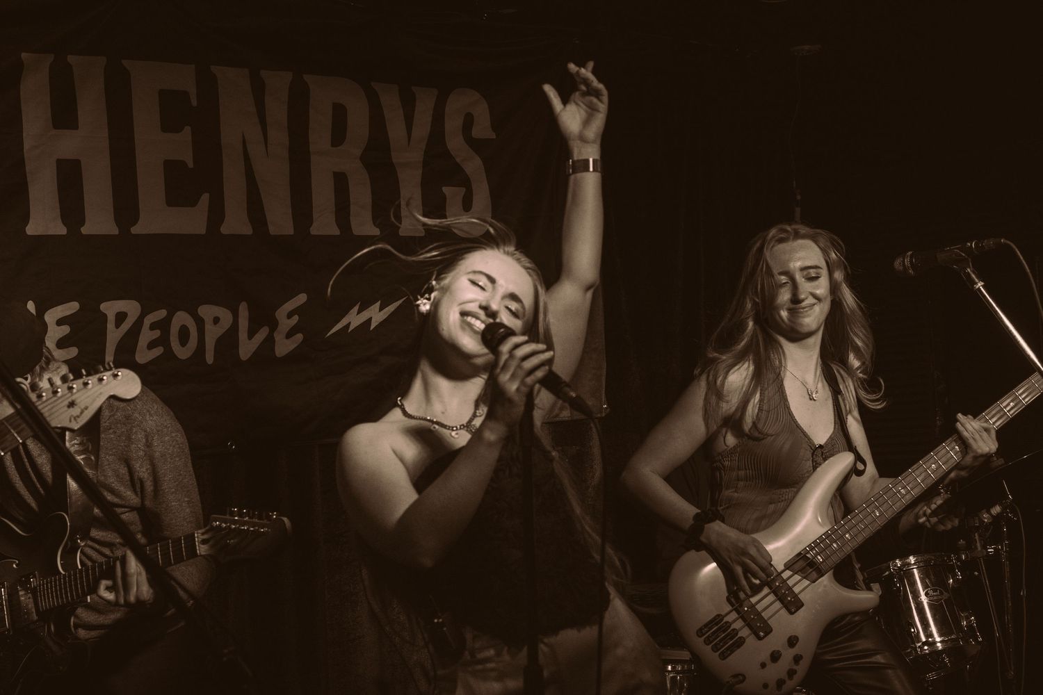 Female vocalist performs passionately on stage at Henrys bar with band members playing guitar and bass behind her.