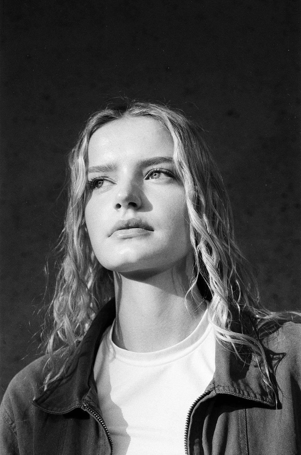 Black and white portrait of a person with wavy blonde hair looking upward while wearing a white shirt and dark jacket.