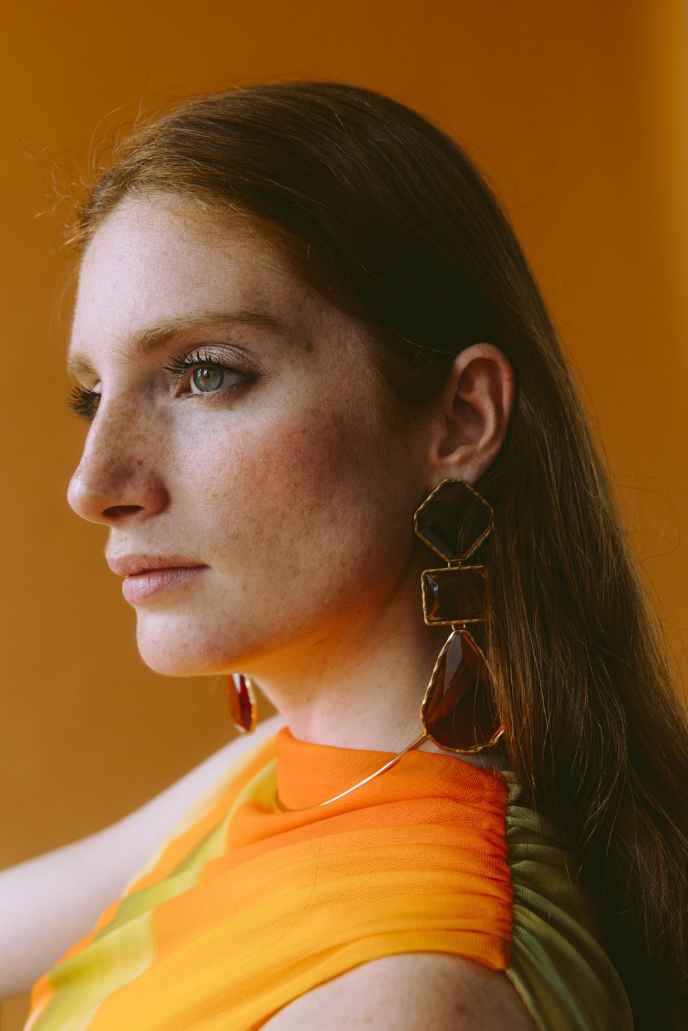 Close-up profile portrait with geometric statement earrings wearing orange top against warm amber background.