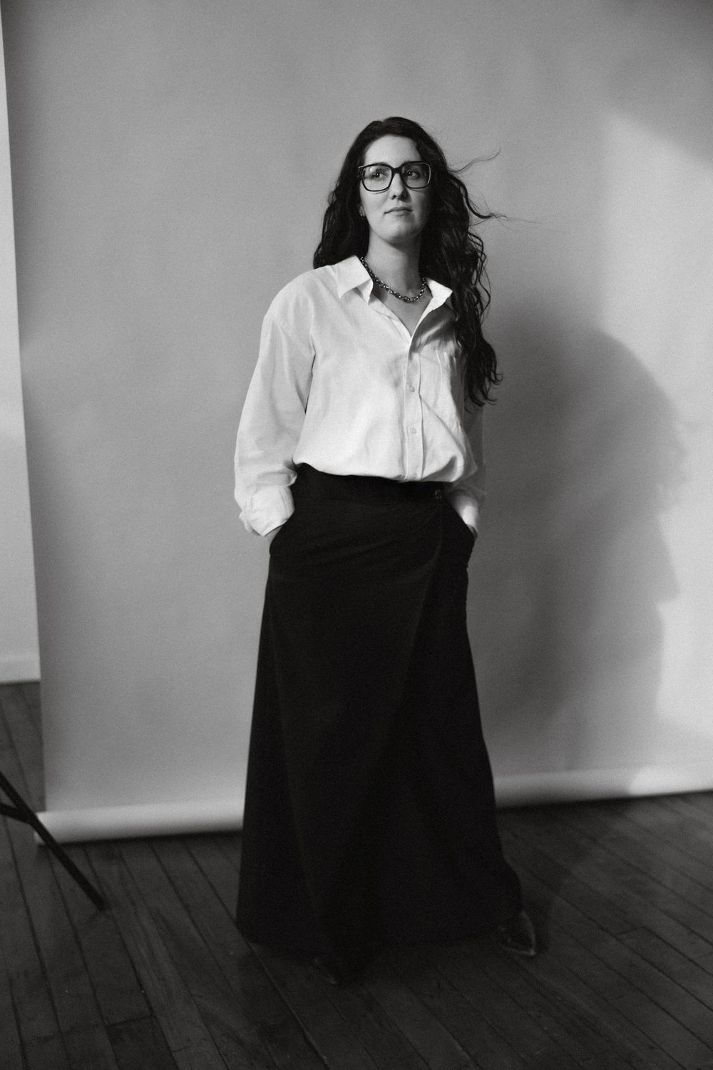 Black and white portrait of someone wearing glasses, a white button-down shirt and long black skirt in a minimalist setting.