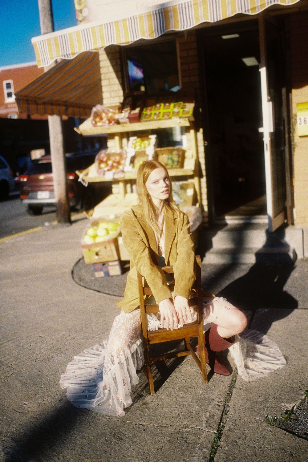 A retro-style street scene with someone in a tan jacket sitting on a wooden chair by a storefront with produce displays.