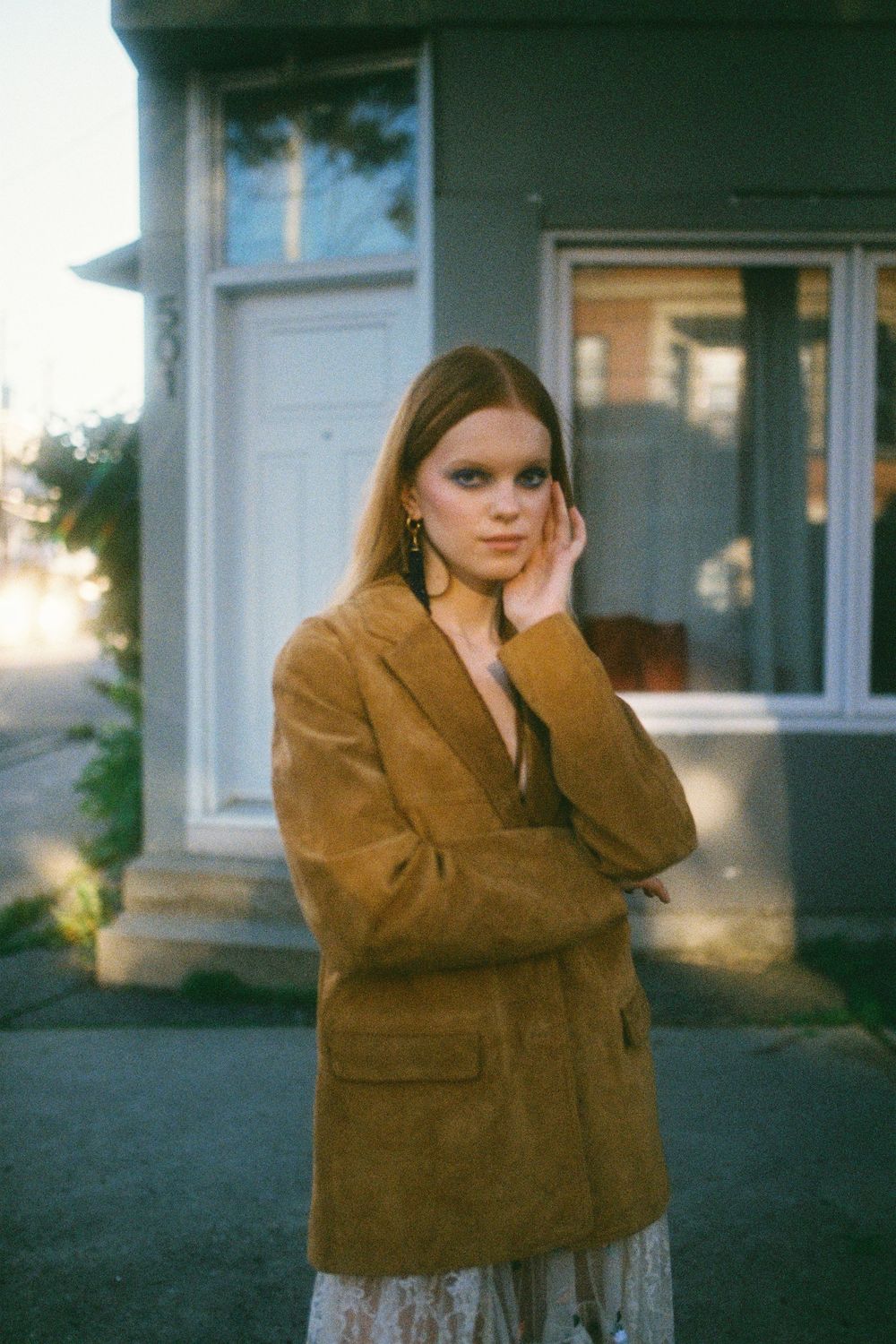 A person in a brown coat stands in front of a building in a contemplative pose at dusk.