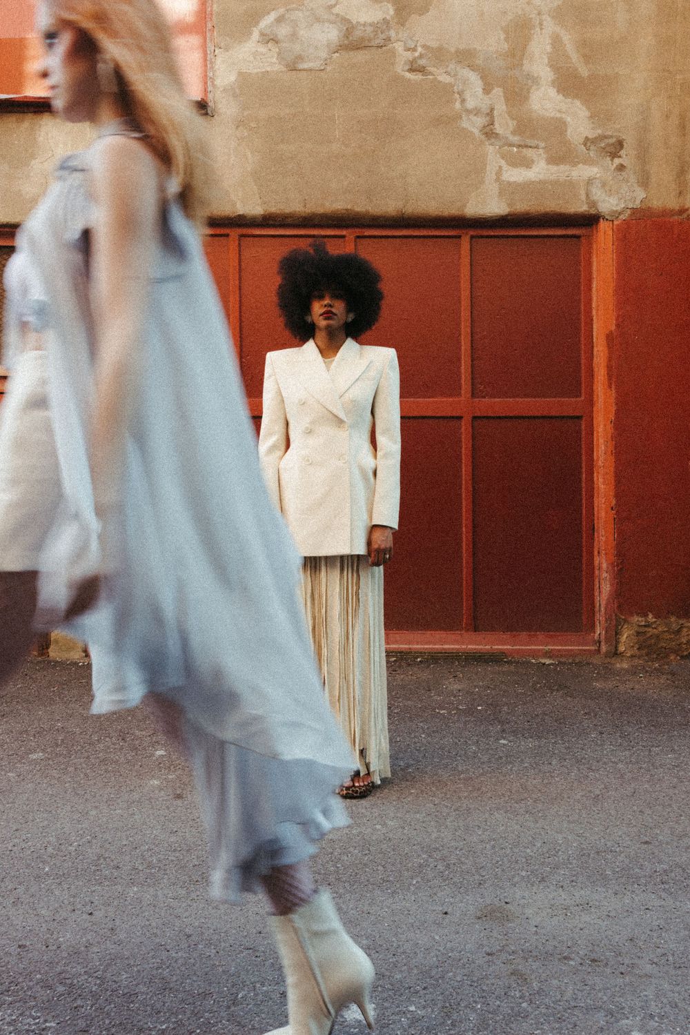 Fashion portrait with person in white blazer and striped pants against rust-colored wall, while blurred figure moves past in motion.