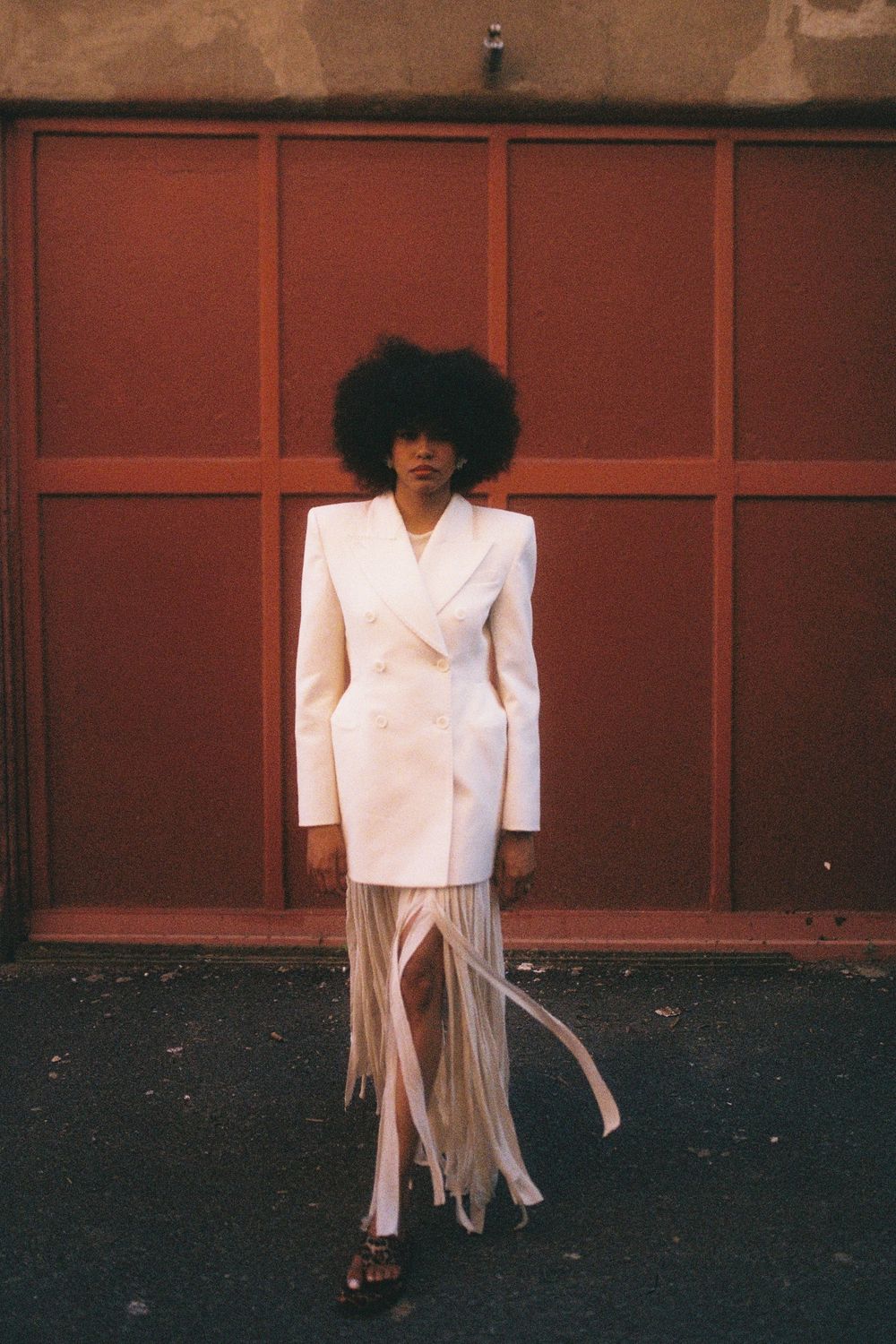 A fashion series showing a person in a white structured jacket and fringe skirt ensemble against a reddish-brown backdrop.