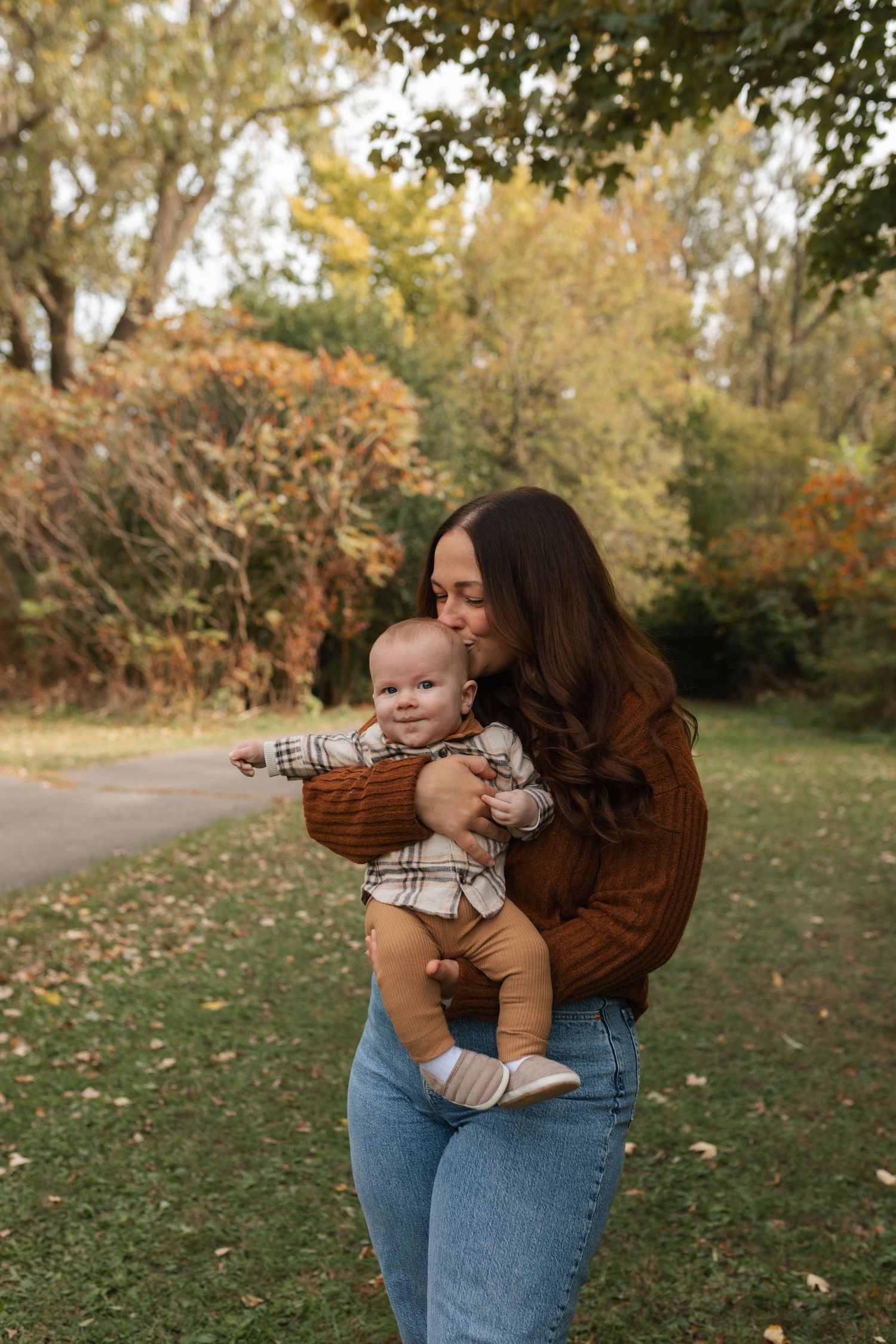 A brunette mother in a brown sweater kisses her smiling baby while standing in a colorful autumn park.