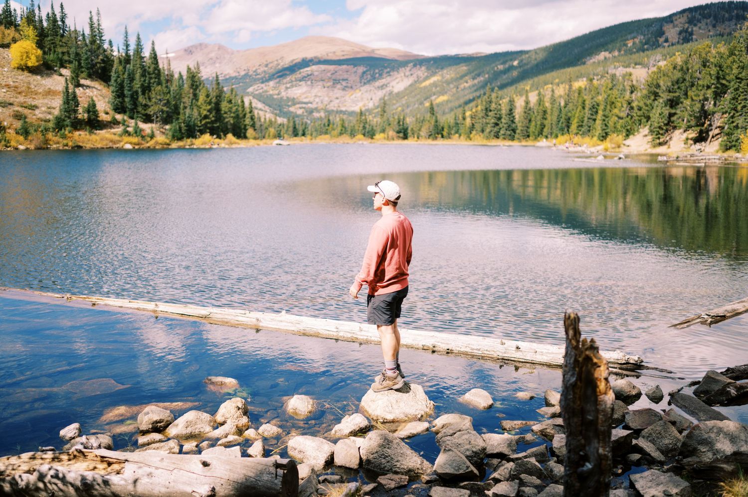 Someone stands on a rock overlooking a pristine mountain lake surrounded by evergreen forest and rocky peaks.