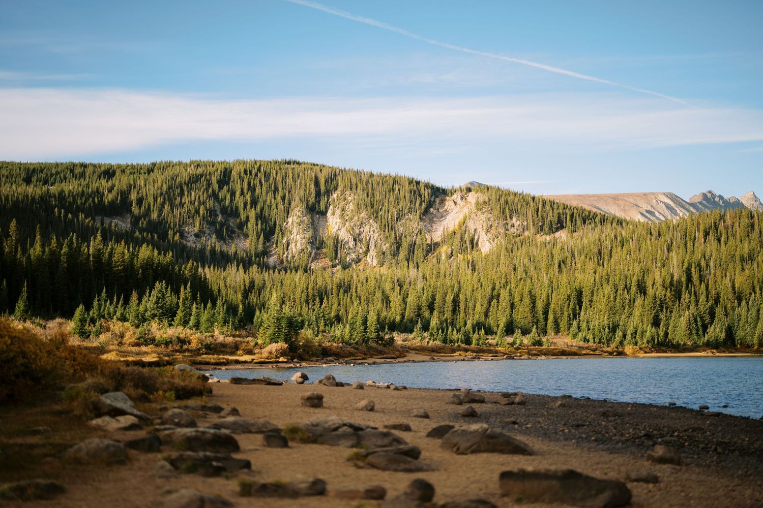 A scenic mountain lake surrounded by dense pine forest and rocky outcrops under blue sky with scattered clouds.