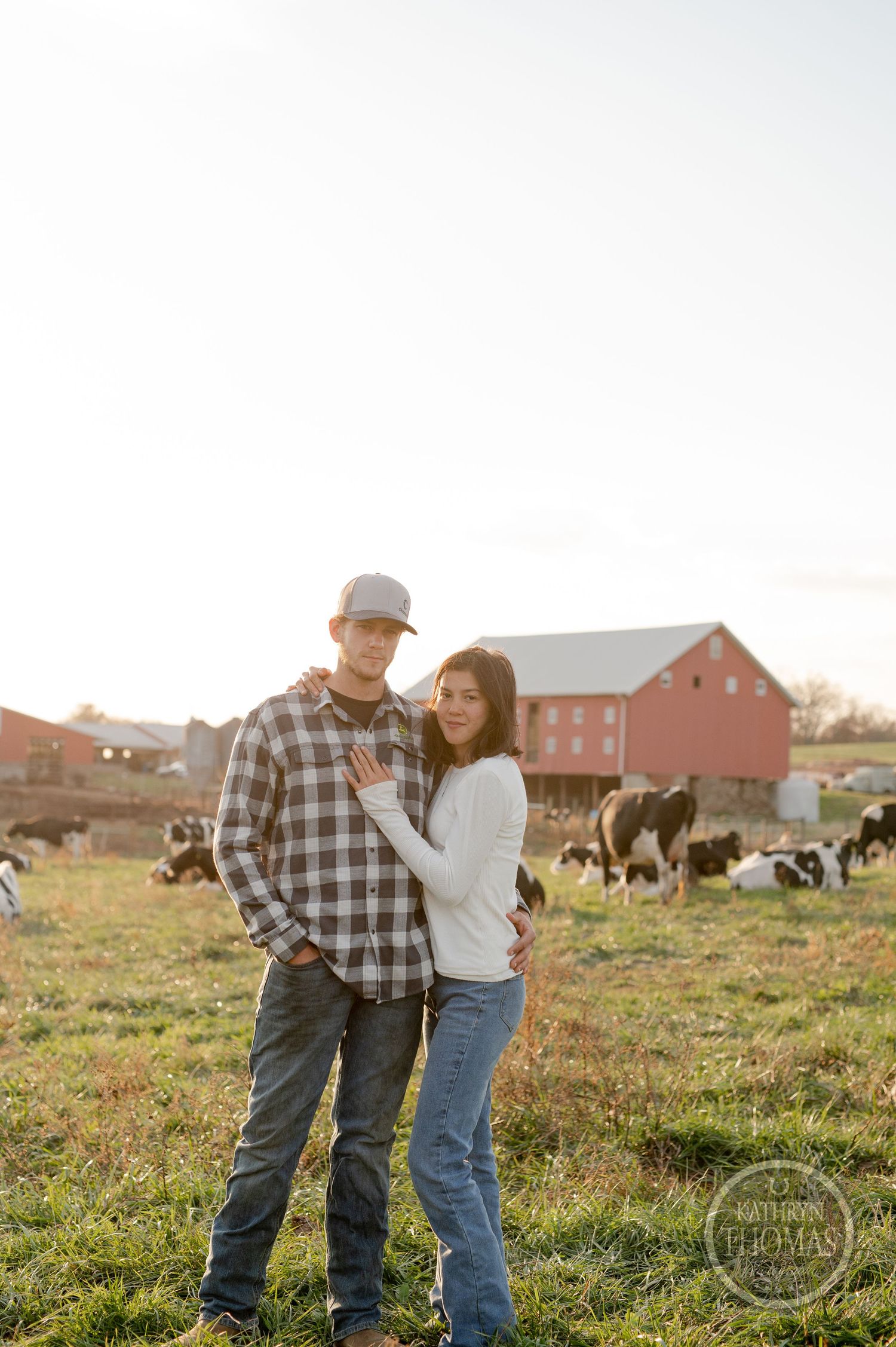 A couple poses together on their farm with grazing cattle and a red barn in the background.