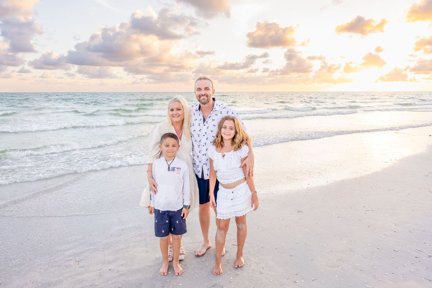Happy family of four poses together on a white sandy beach during a beautiful golden sunset.
